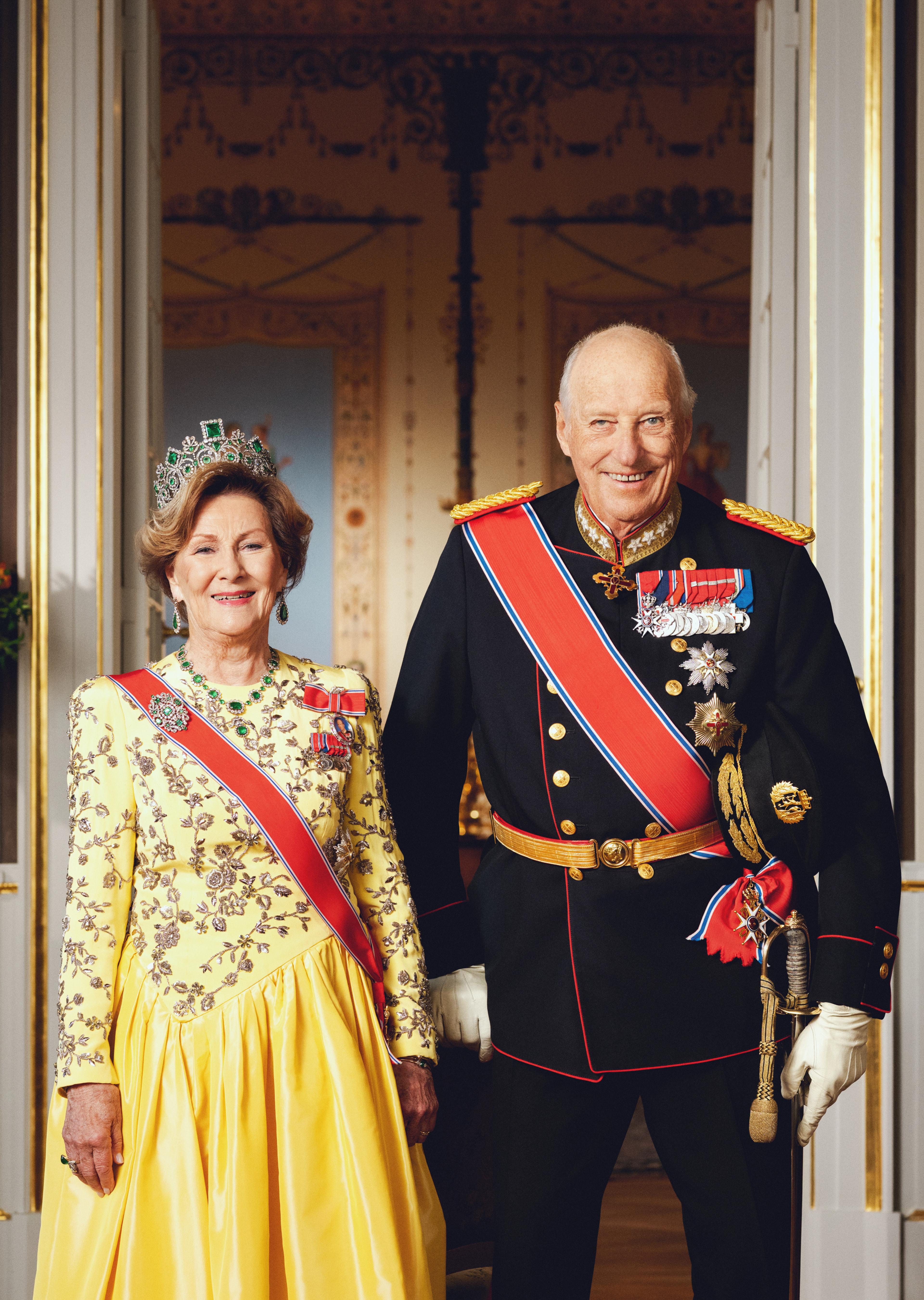Kongeparet står smilende - Kongen i mørk gallauniform med ordensbånd og medaljer, Dronningen i gul sid, gul kjole med ordener og tiara.