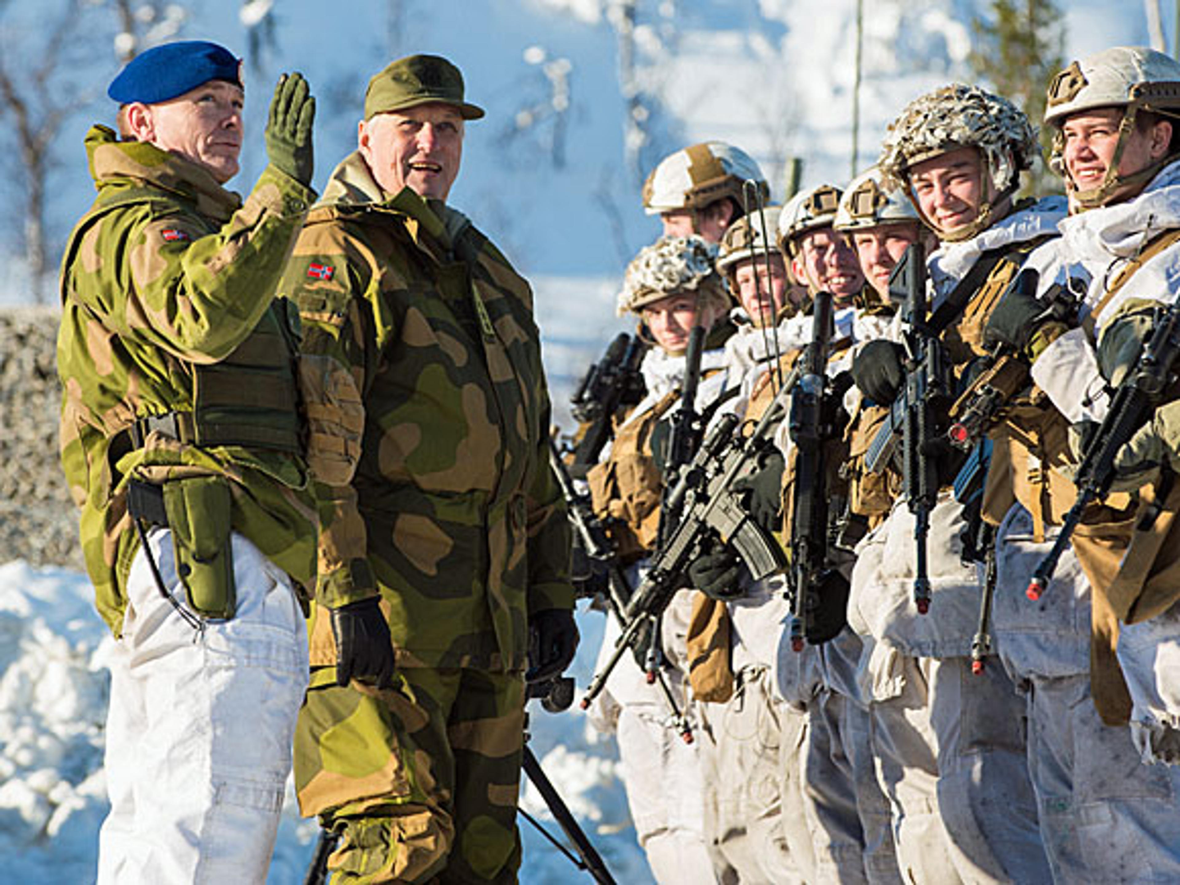 Kong Harald i feltuniform lytter til brigadesjefen. Menige soldater i vinteruniformer står foran dem.