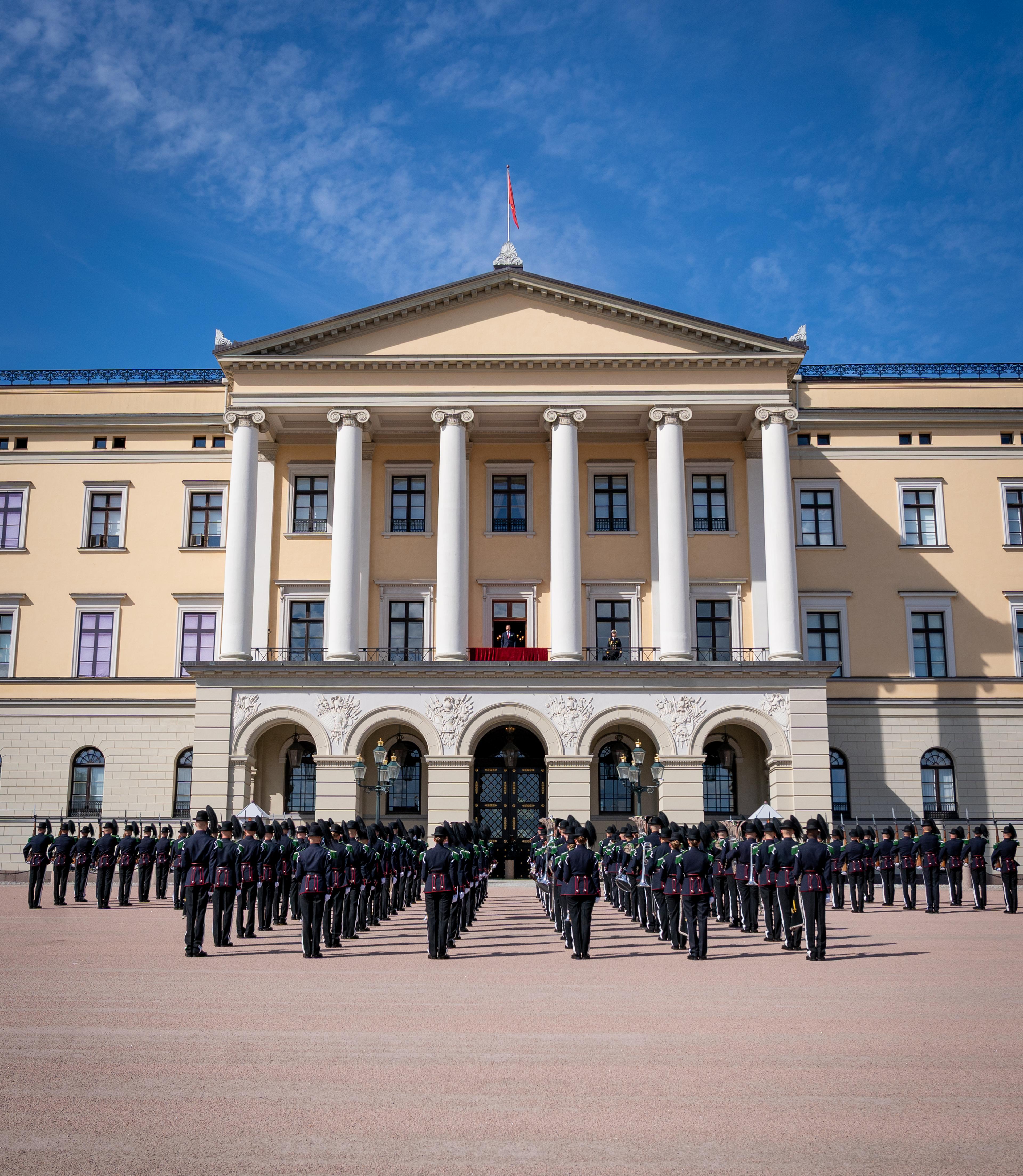 Musikk- og Drilltroppen står oppstilt foran Slottet. På Slottsbalkongen står Kronprins Haakon.