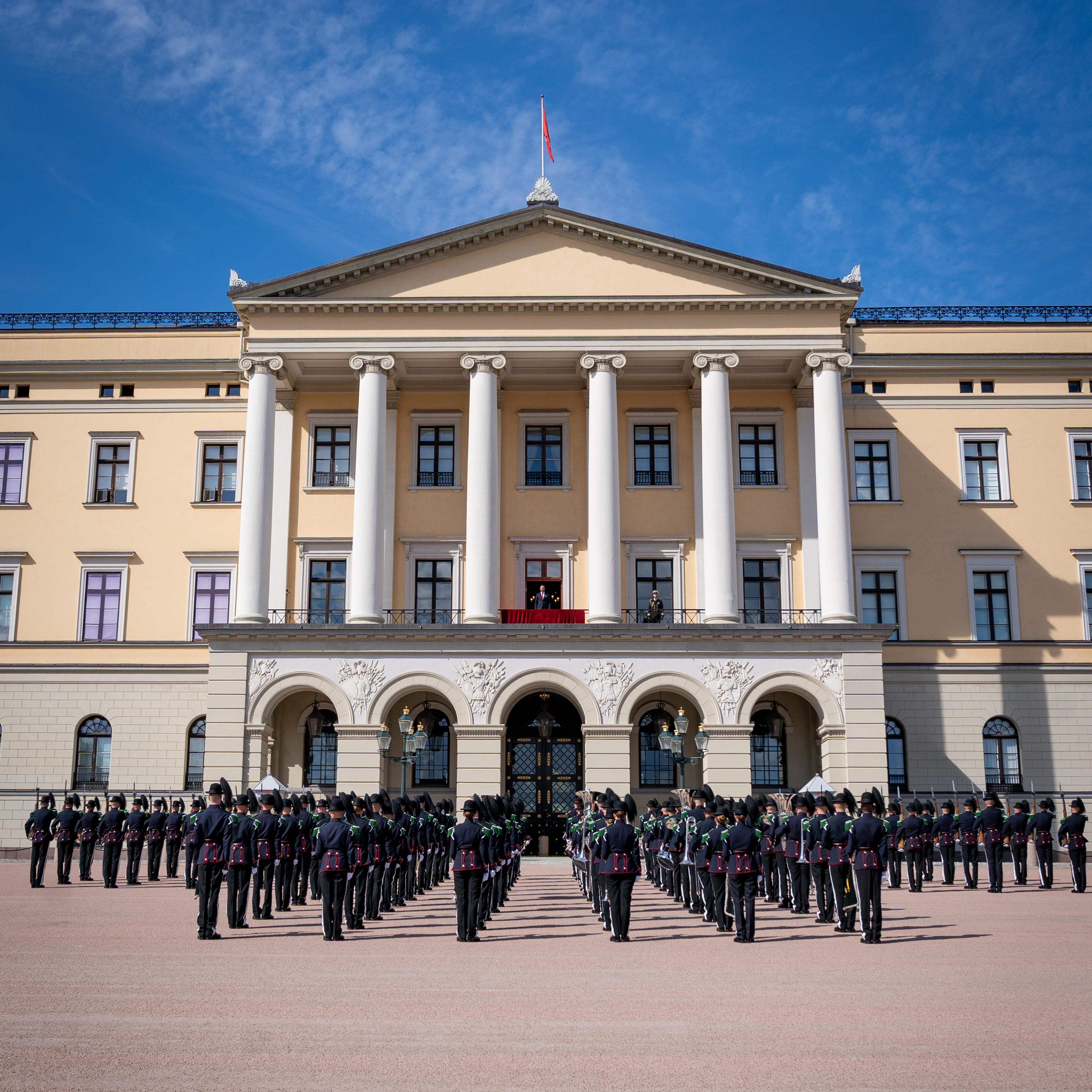 Musikk- og Drilltroppen står oppstilt foran Slottet. På Slottsbalkongen står Kronprins Haakon.
