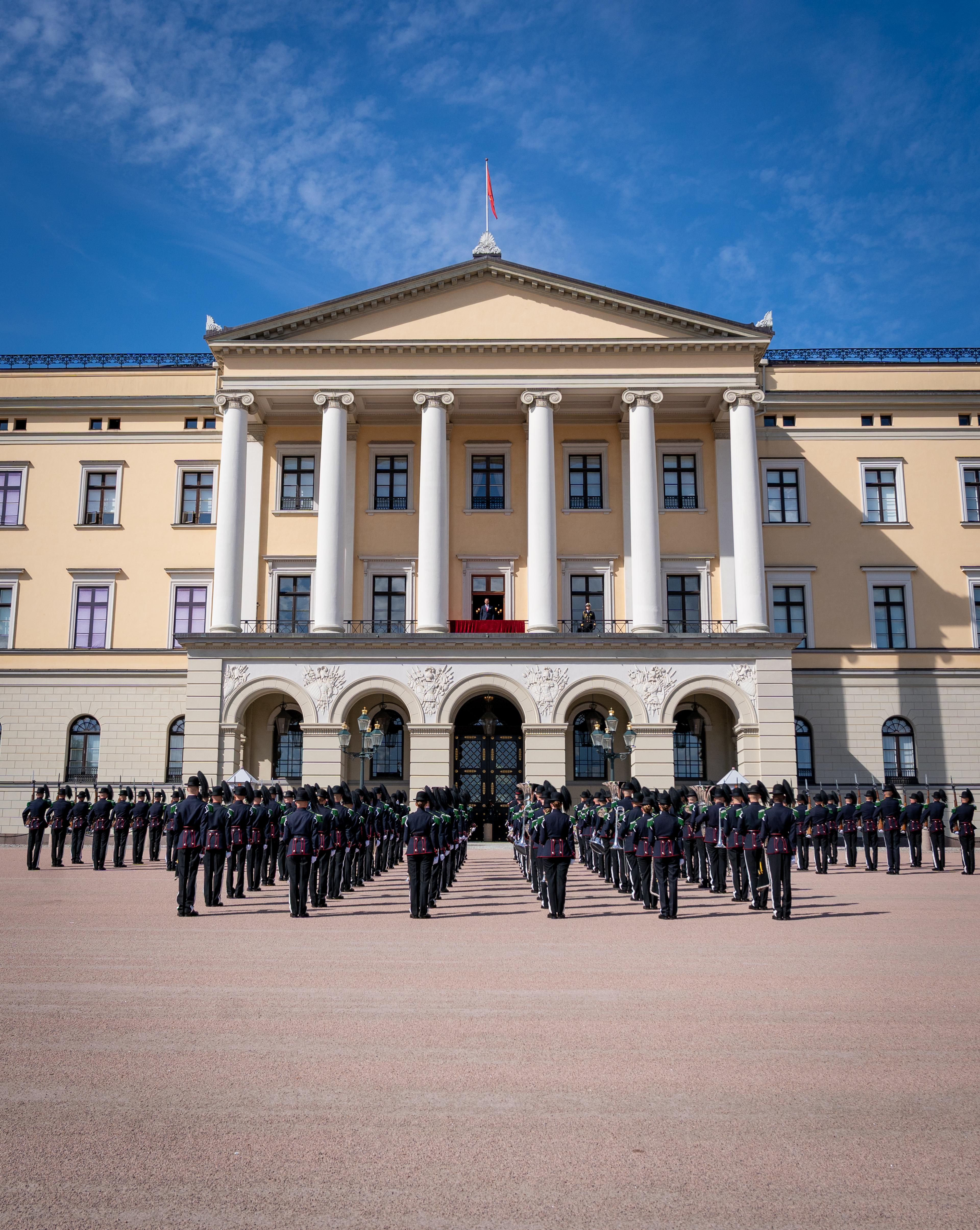Musikk- og Drilltroppen står oppstilt foran Slottet. På Slottsbalkongen står Kronprins Haakon.