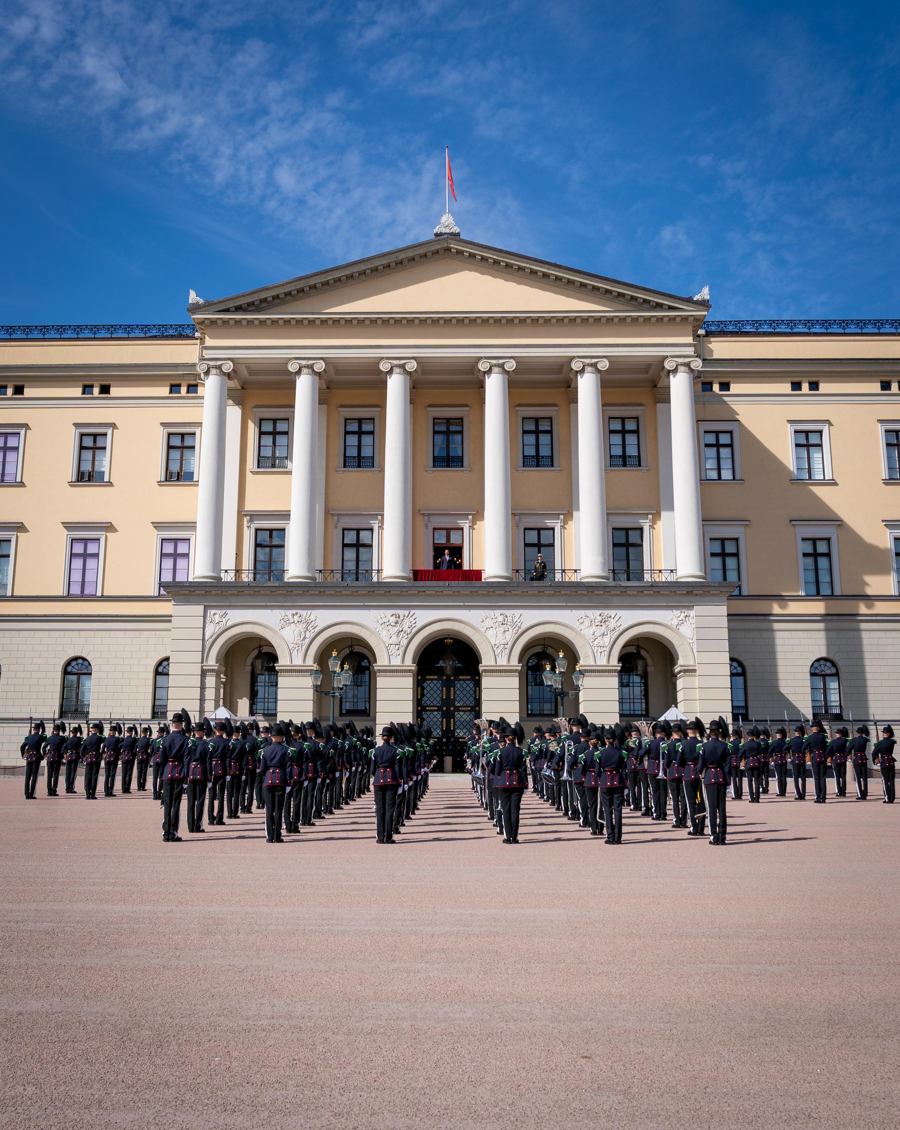 Musikk- og Drilltroppen står oppstilt foran Slottet. På Slottsbalkongen står Kronprins Haakon.