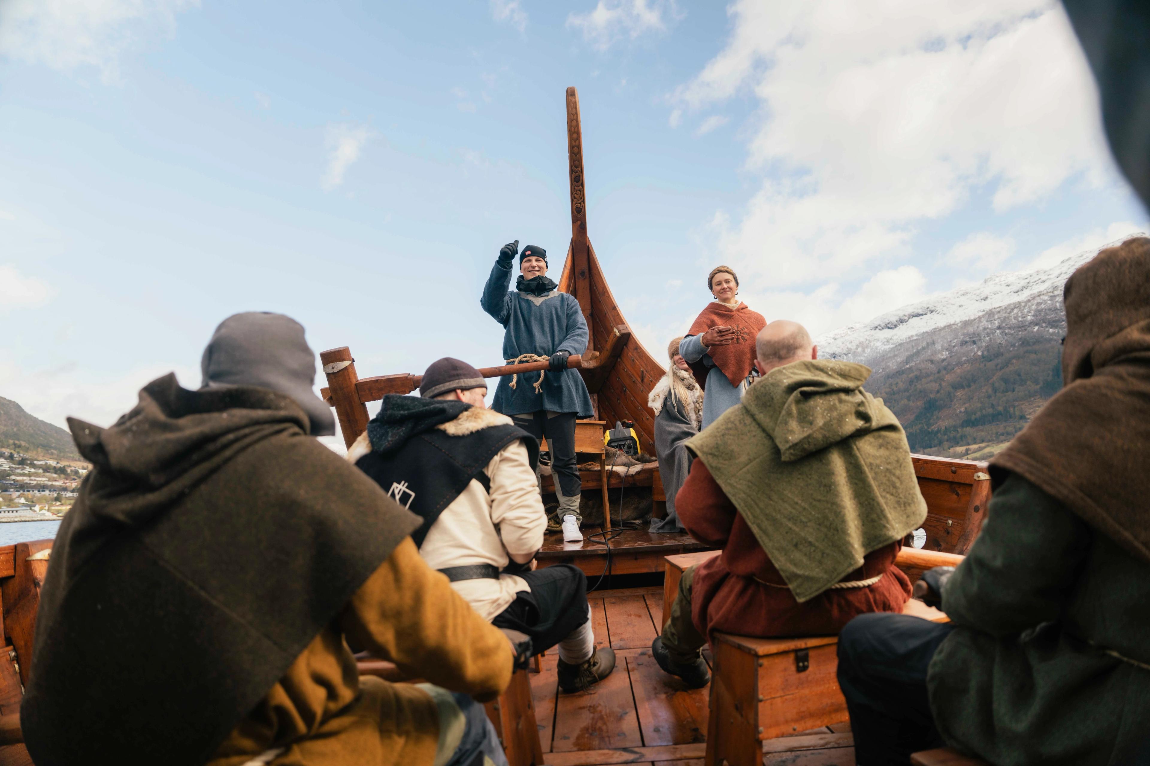 Prins Sverre Magnus står bakerst i et vikingskip, med en hånd på roret og den andre som peker i været. I forgrunnen sitter flere personer kledd i vikingklær med hver sin åre. I bakgrunnen er fjell og blå himmel i sikte
