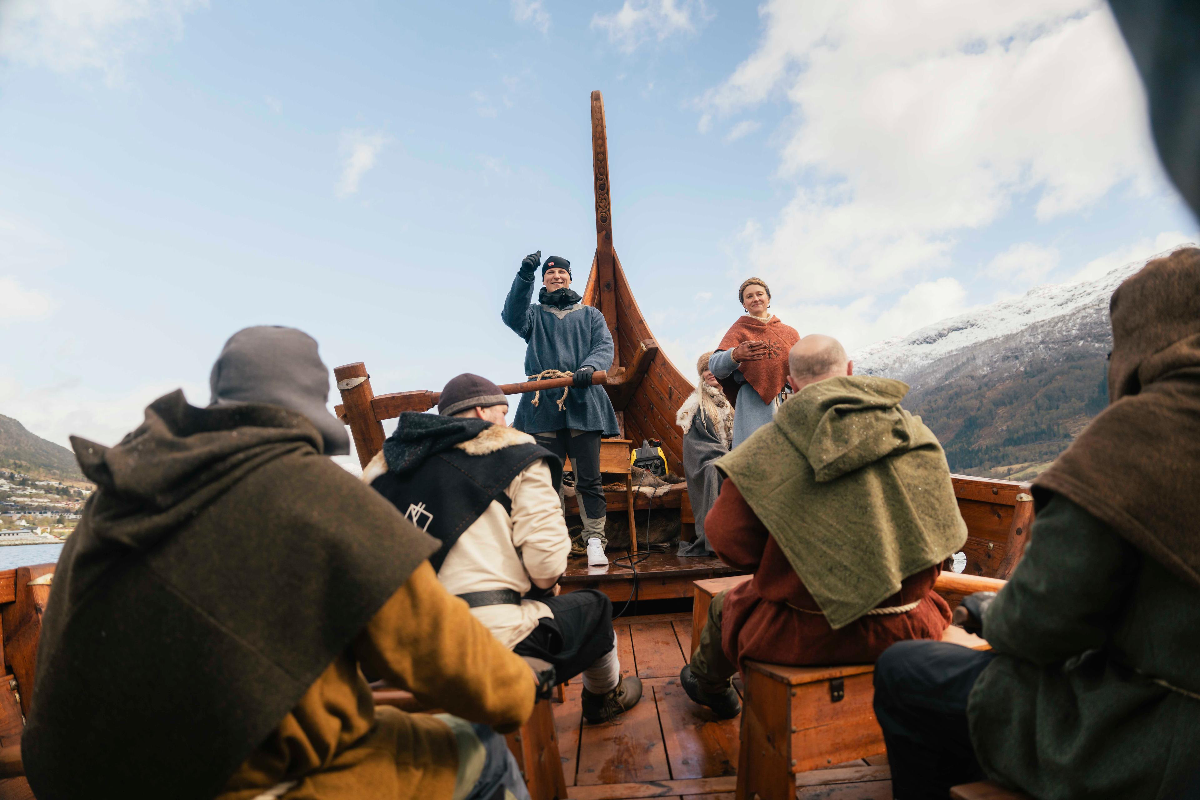 Prins Sverre Magnus står bakerst i et vikingskip, med en hånd på roret og den andre som peker i været. I forgrunnen sitter flere personer kledd i vikingklær med hver sin åre. I bakgrunnen er fjell og blå himmel i sikte