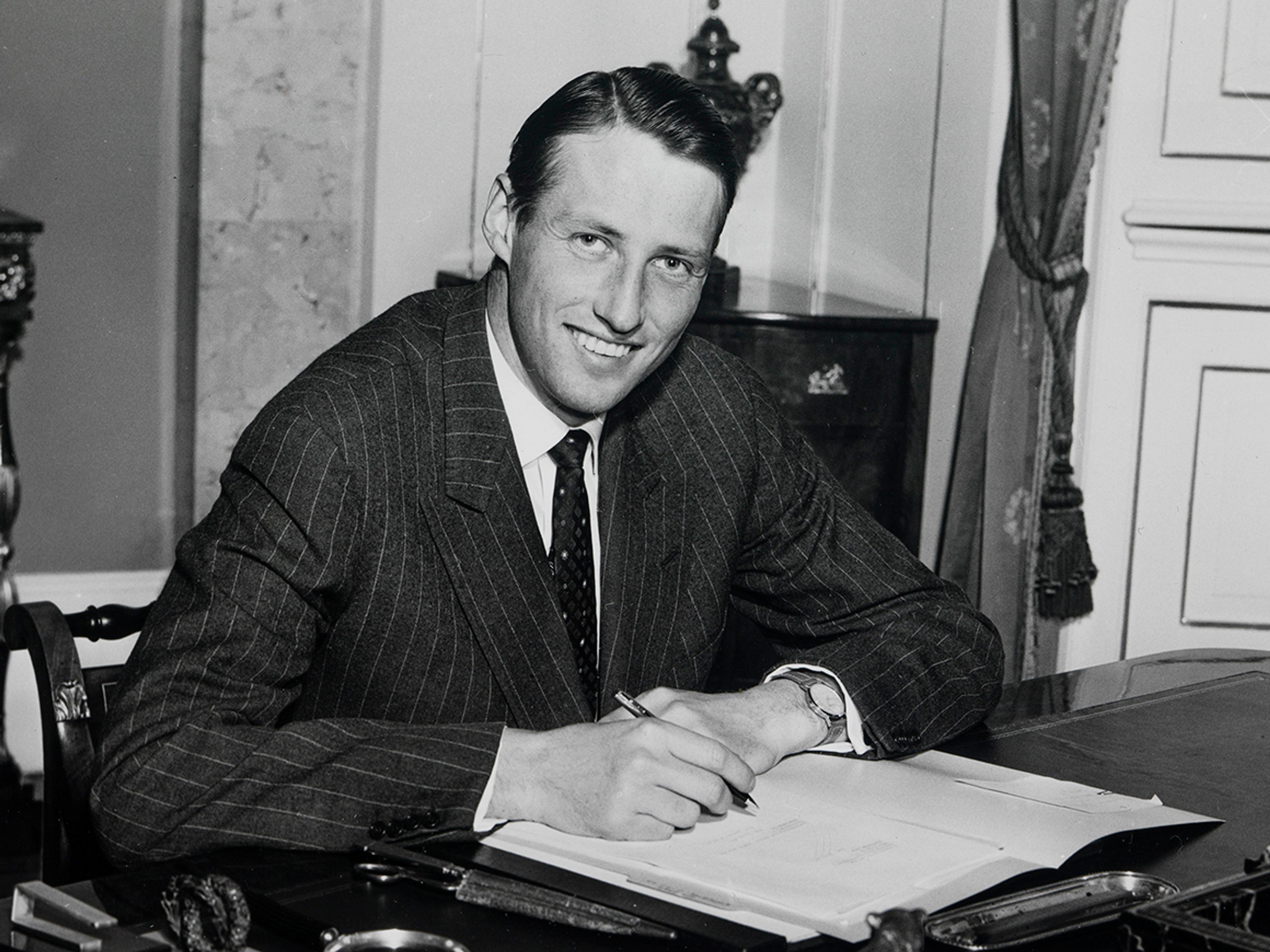 Sort-hvitt. Kronprinsen sitter bak skrivebordet sitt og ser smilende mot kameraet.