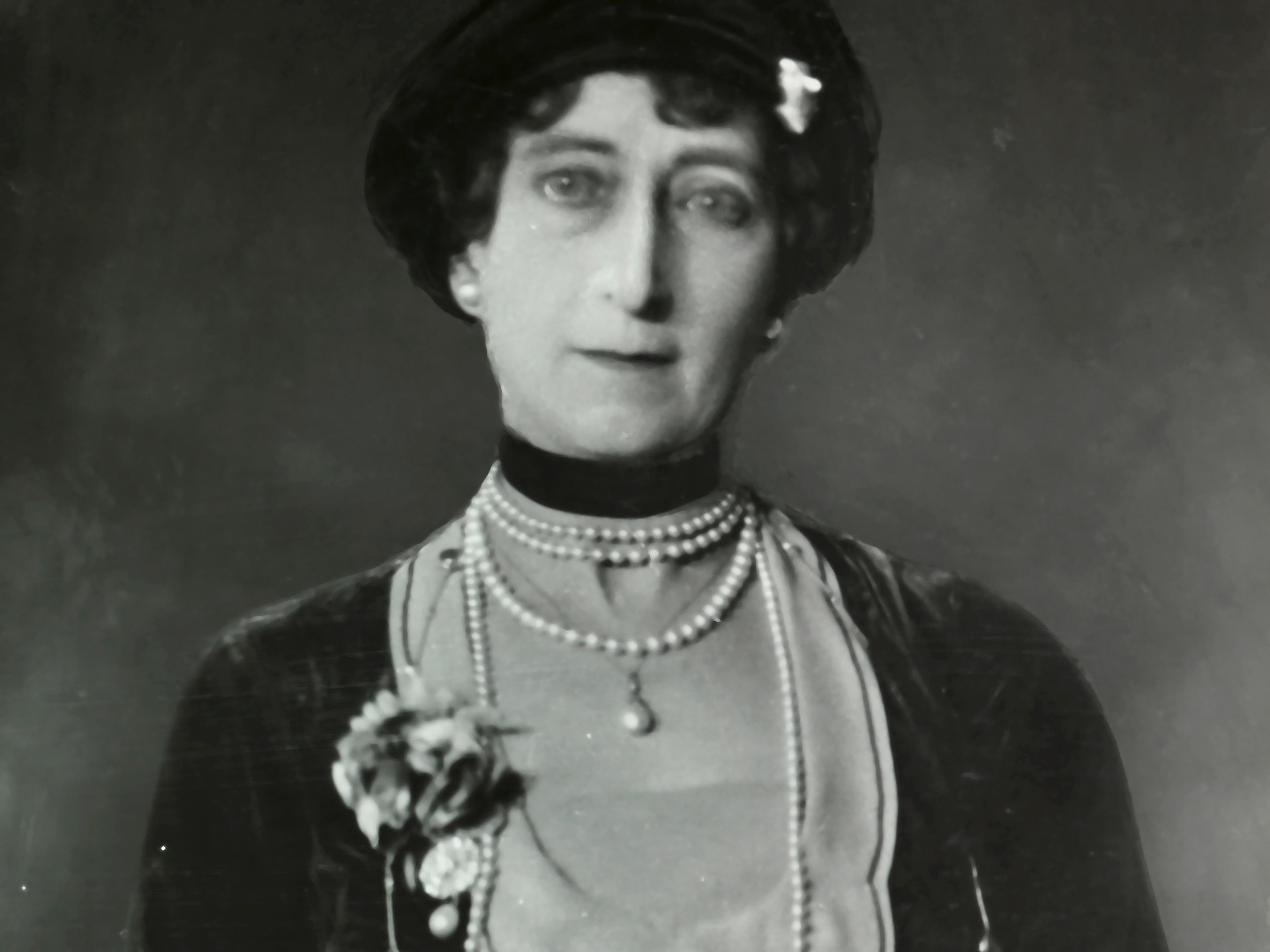 Portrett av Dronning Maud med langt perlekjede og muffe i skinn.