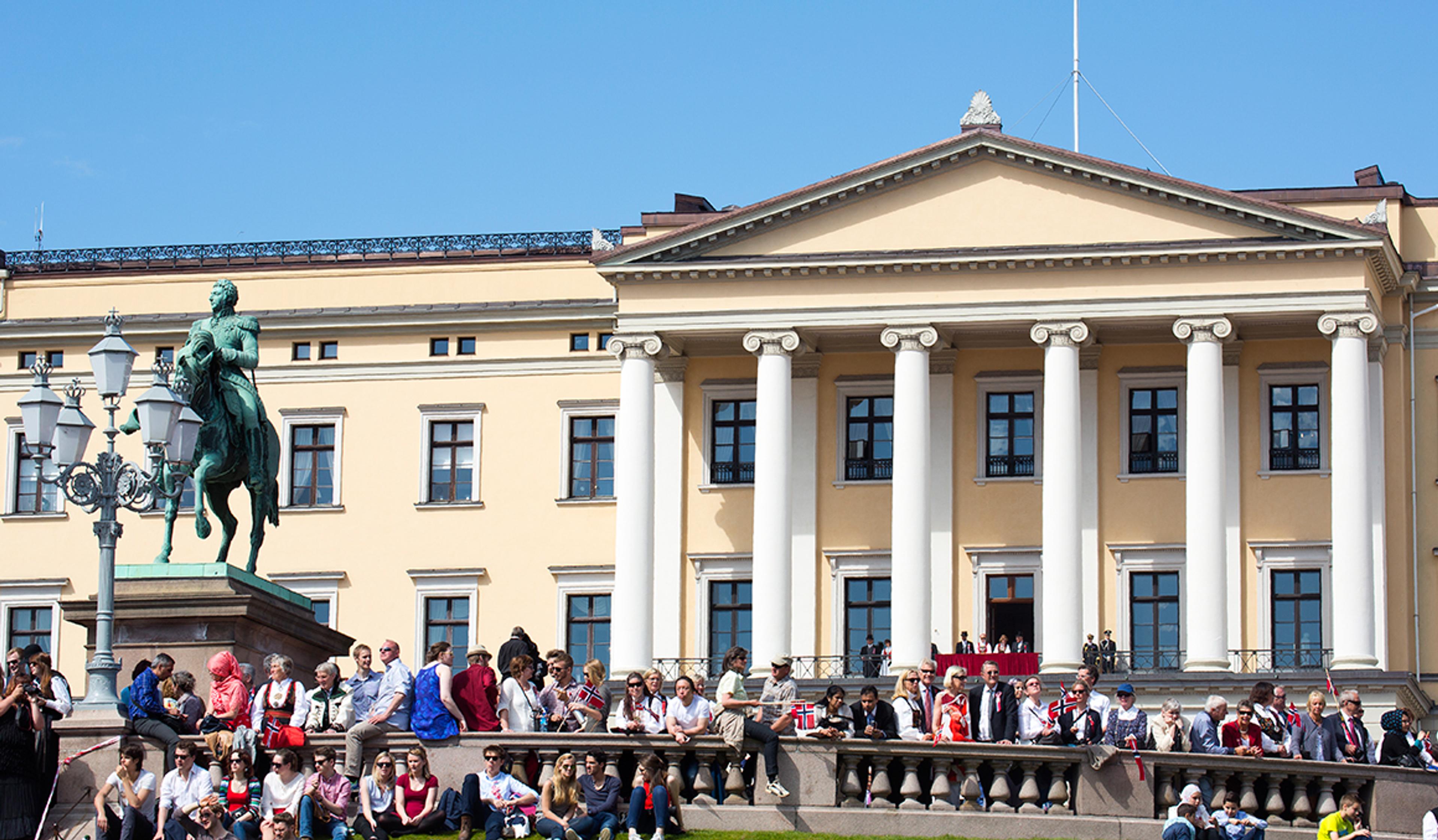 Utsnitt av Slottets fasade med søylefronten og statuen av Kong Carl Johan i forgrunnen. Det er kongelige på Slottsbalkongen og mange festkledde mennesker langs balustraden utenfor.