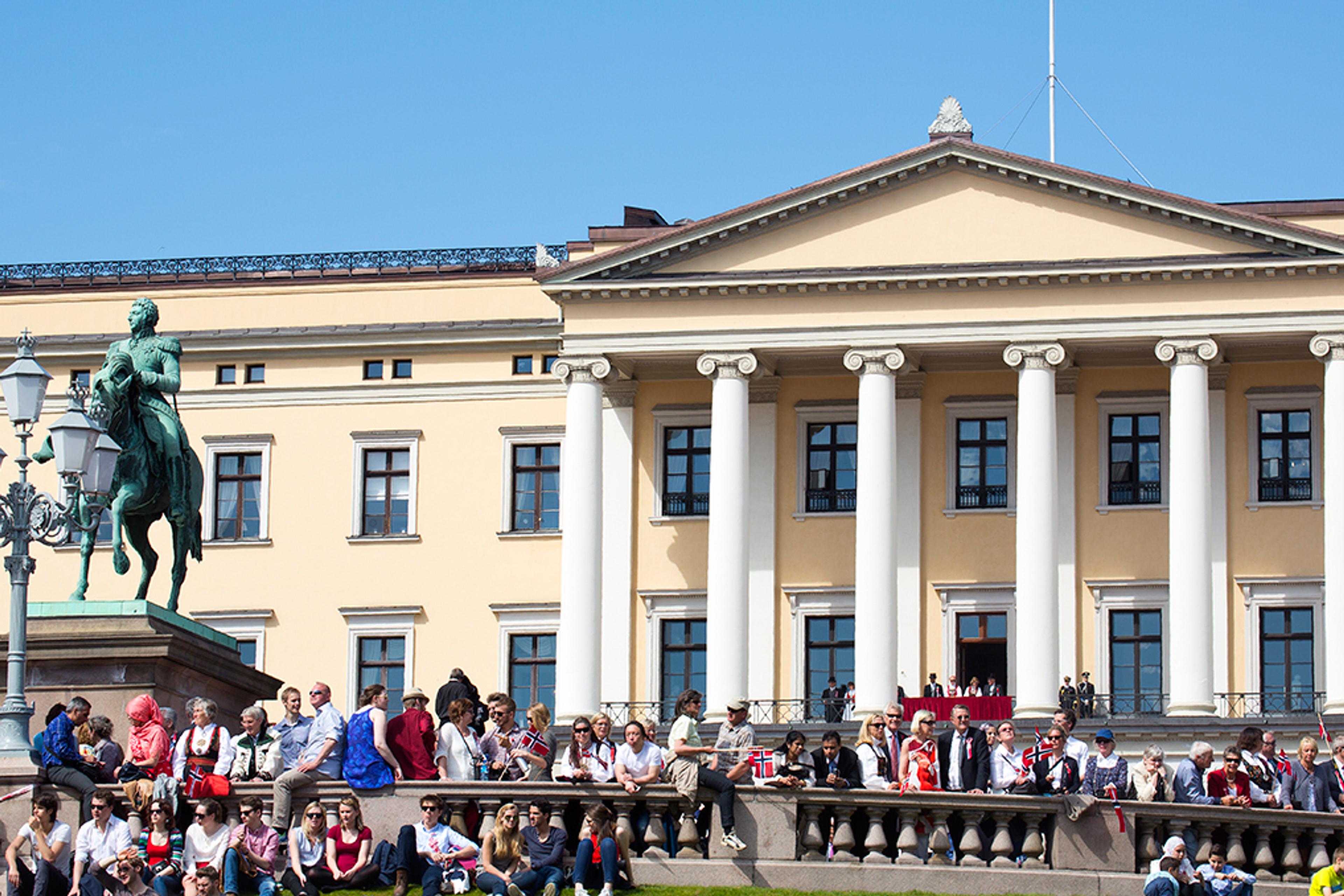 Utsnitt av Slottets fasade med søylefronten og statuen av Kong Carl Johan i forgrunnen. Det er kongelige på Slottsbalkongen og mange festkledde mennesker langs balustraden utenfor.