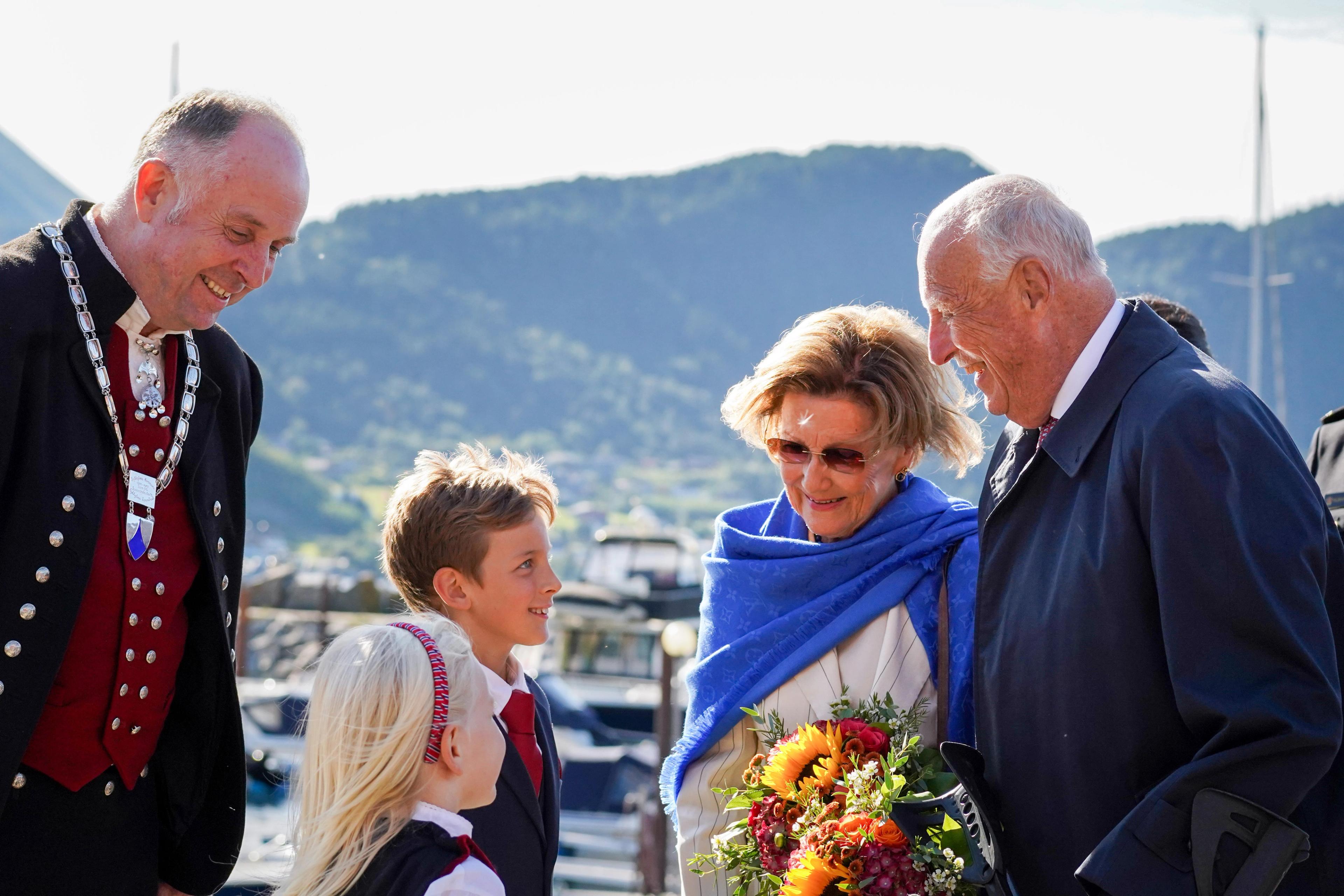 Kong Harald og Viljar ser smilende på hverandre. Dronning Sonja ser smilende på Martine, med en ordfører i bunad støttende bak.