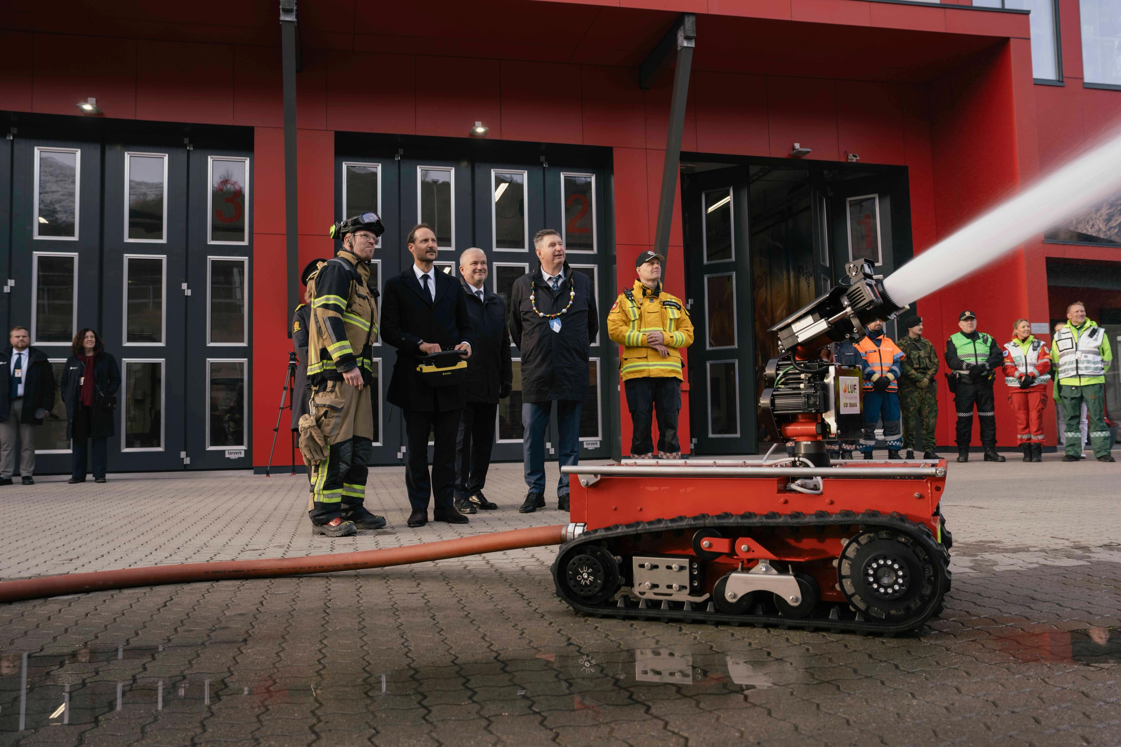 Kong Haakon står med fjernkontroll og styrer en brannvernrød robot på beltehjul som spruter vann ut av bildet.