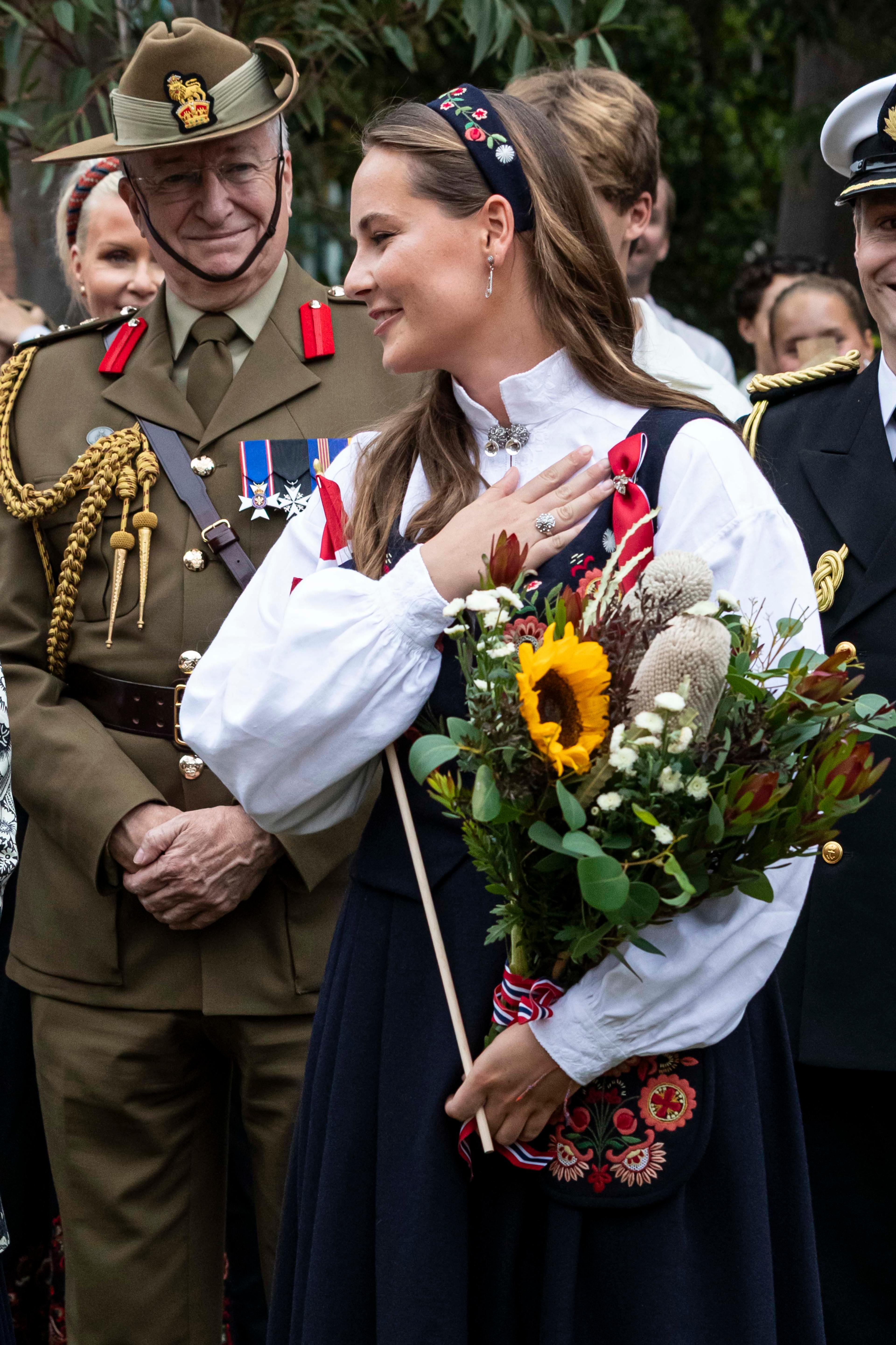 Prinsessen står smilende med en blomsterbukett og et norsk i armene. Hun er kledd i bunad og har lagt hånden over hjertet.