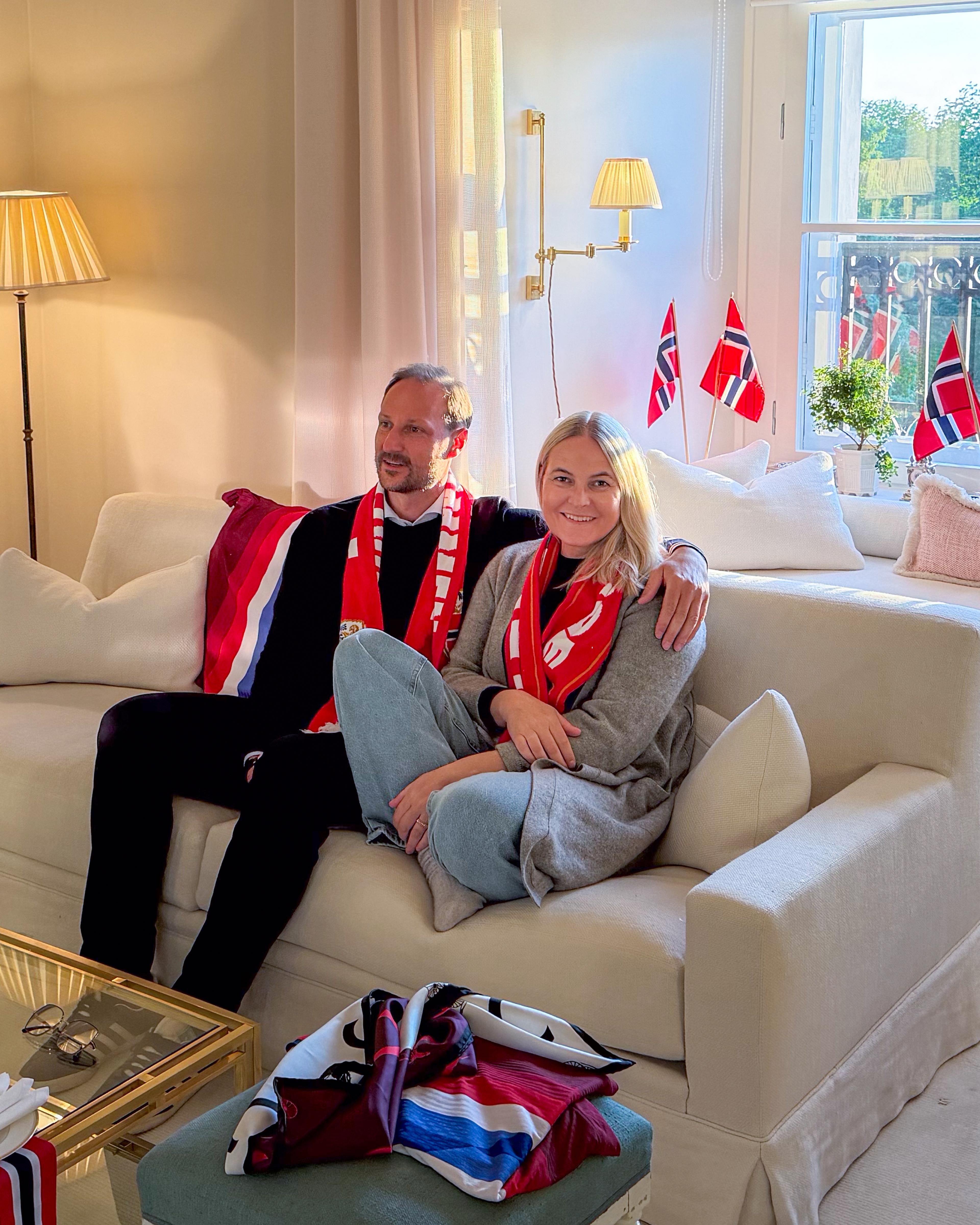 Kronprinsparet sitter i sofaen med norsk fotballskjerf rundt halsen, Kronprinsessen med beina under seg. Det er norske flagg i vinduskarmen.