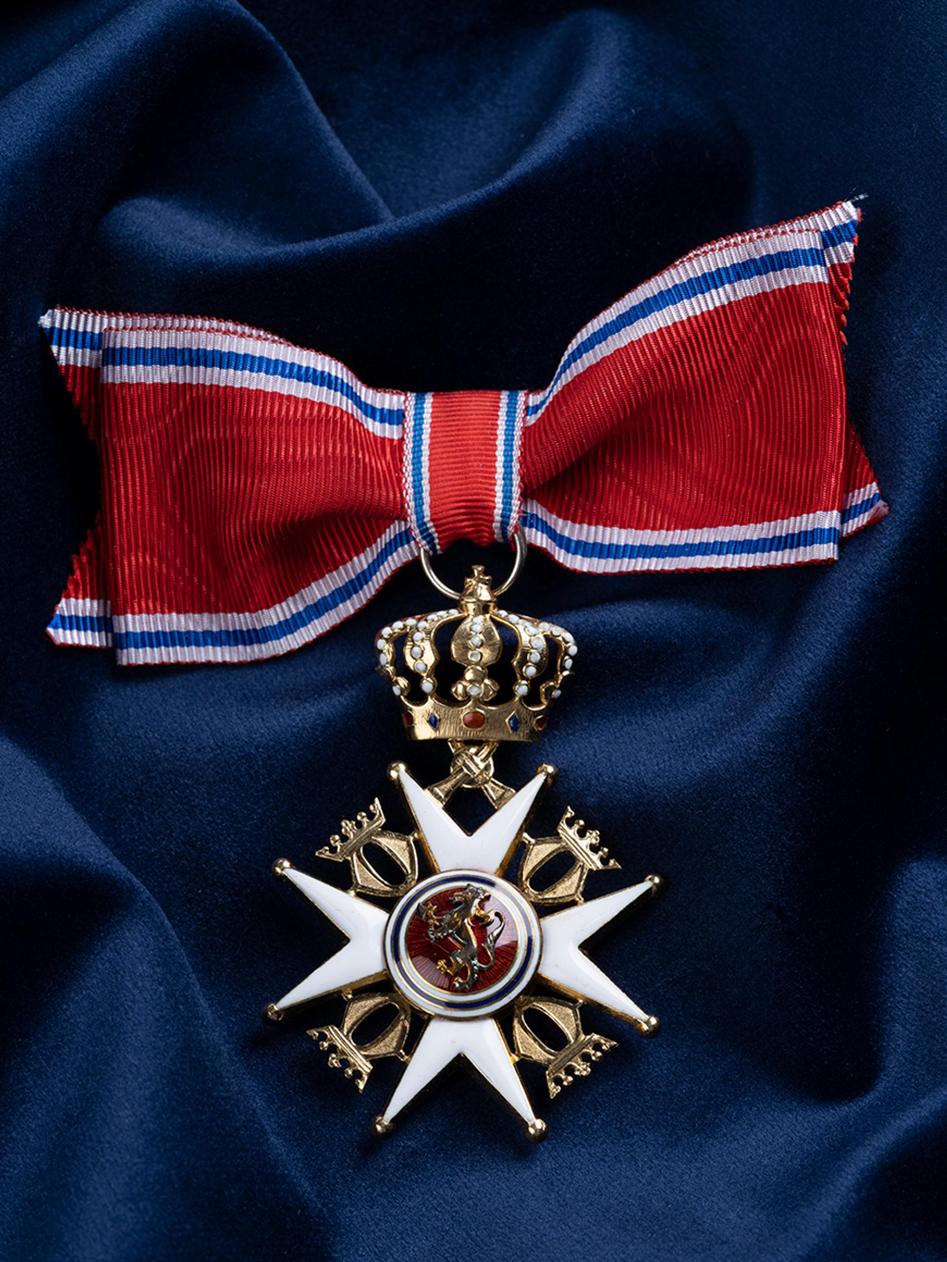 Nærbilde av ordensmerket festet i rødt bånd med blå og hvite striper på en bakgrunn av blått klede.