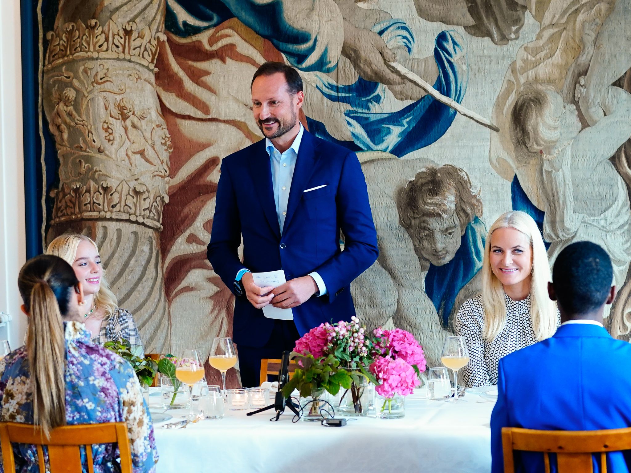 Kronprins Haakon står ved et dekket bord med talekort i hendene og snakker til gjestene rundt bordet. Kronprinsessen sitter ved siden av ham.
