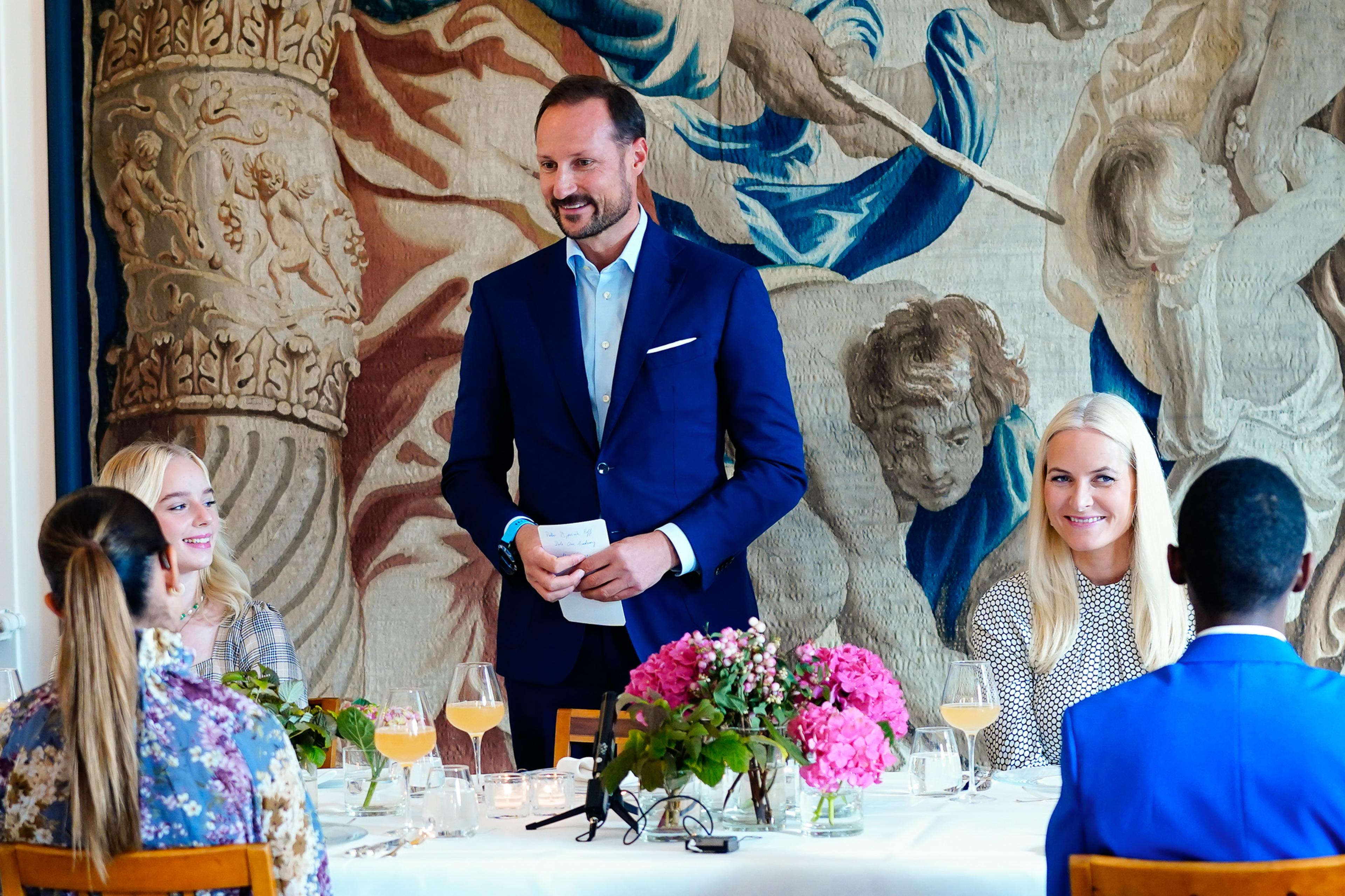 Kronprins Haakon står ved et dekket bord med talekort i hendene og snakker til gjestene rundt bordet. Kronprinsessen sitter ved siden av ham.