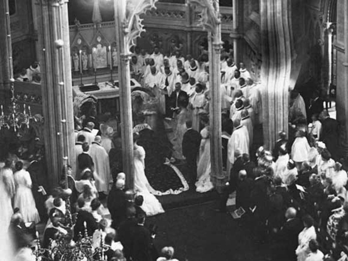 Sort-hvittbilde tatt delvis ovenfra viser Kong Haakon knelende i kroningskappen foran høyalteret omgitt av geistlige. 