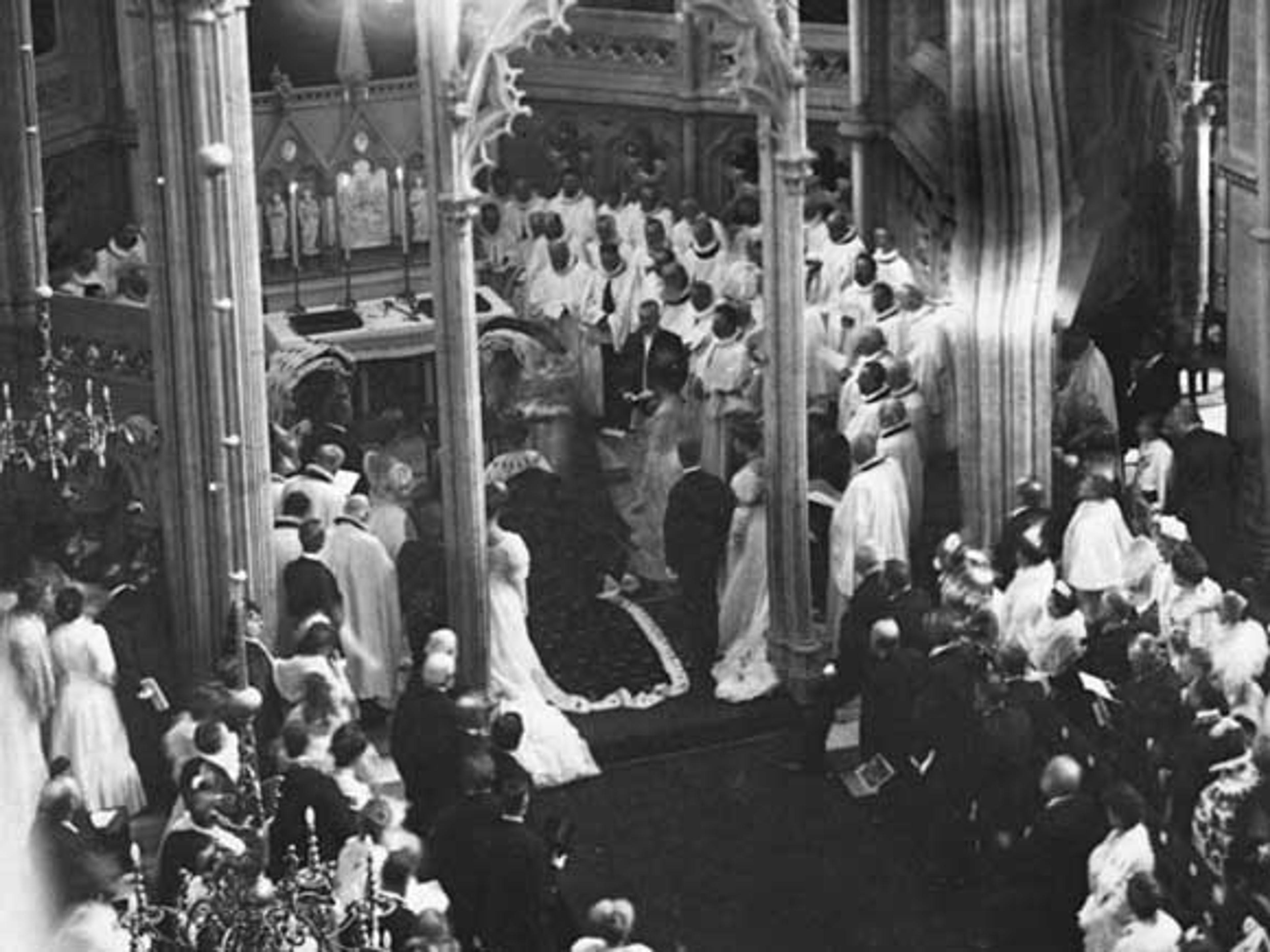 Sort-hvittbilde tatt delvis ovenfra viser Kong Haakon knelende i kroningskappen foran høyalteret omgitt av geistlige.