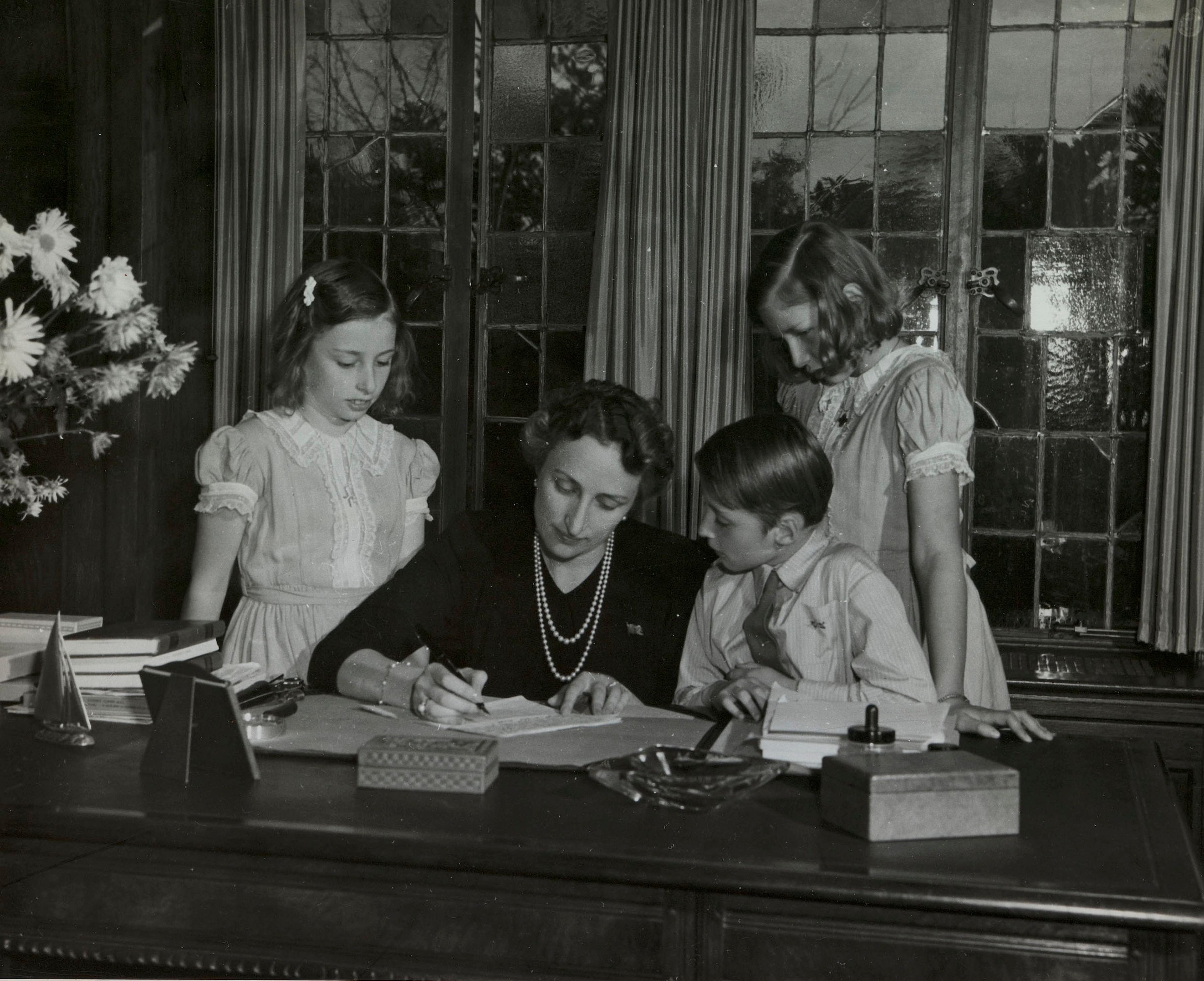 Kronprinsessen sitter ved skrivebordet. Barna står omkring henne og følger med.