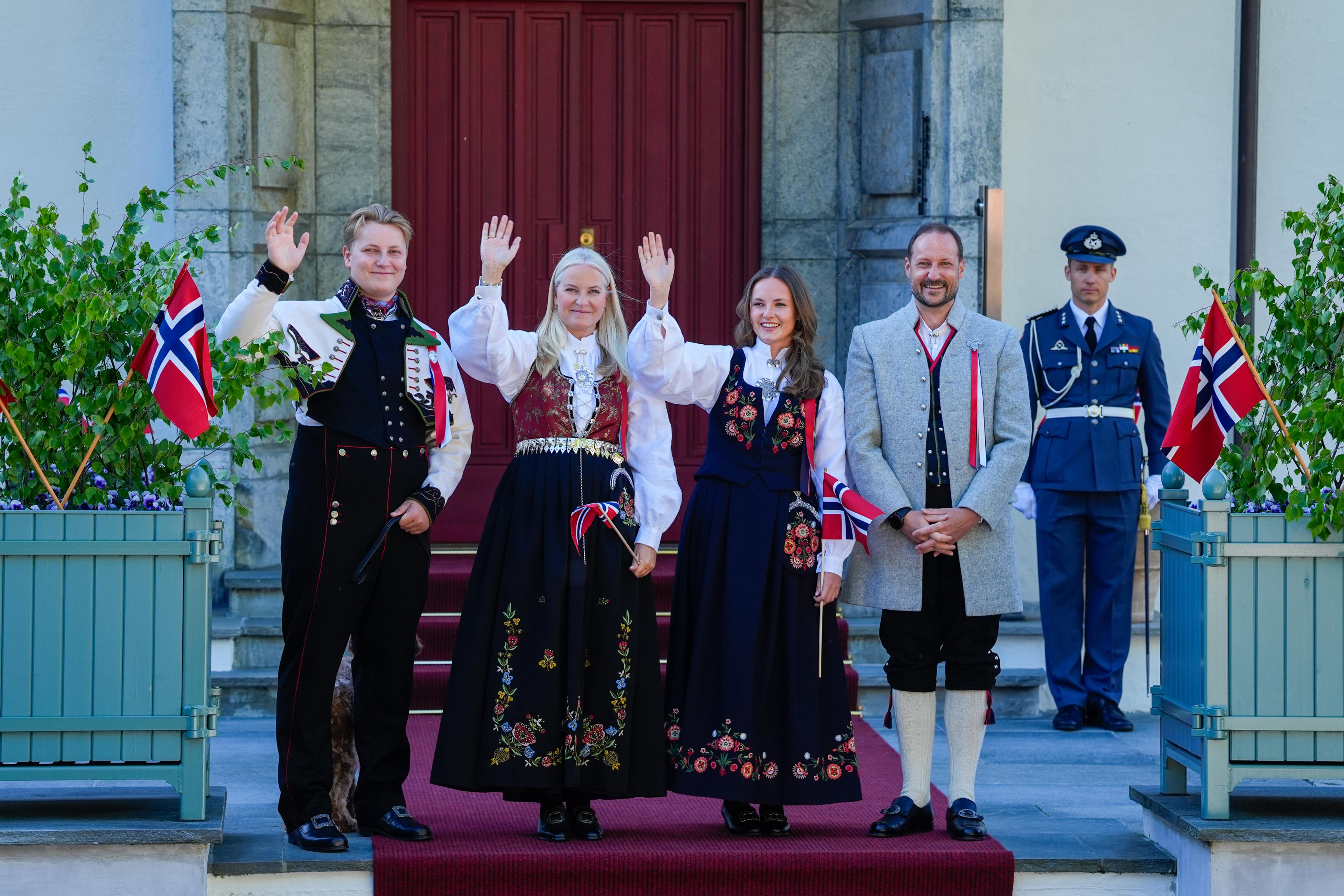 Kronprinsfamilien står på trappa foran inngangsdøra og vinker og hilser i bunad.