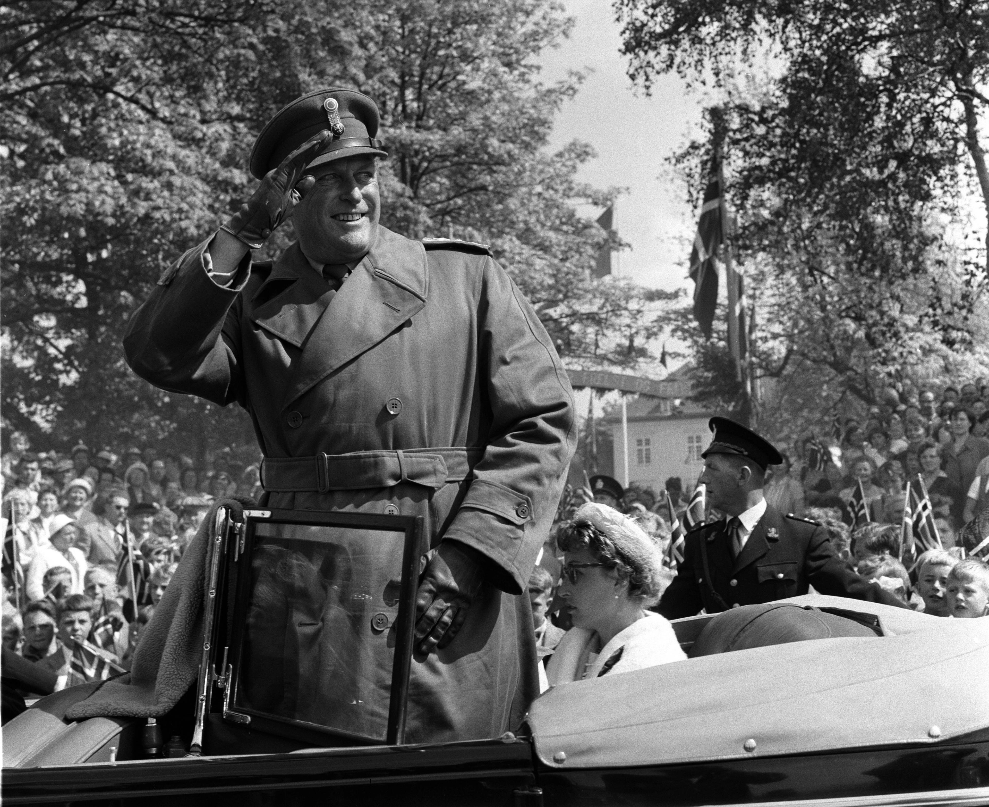 Kongen står smilende oppreist i åpen bil. Han har uniformslue og -frakk på seg og gjør honnør. Det er mange mennesker i bakgrunnen.