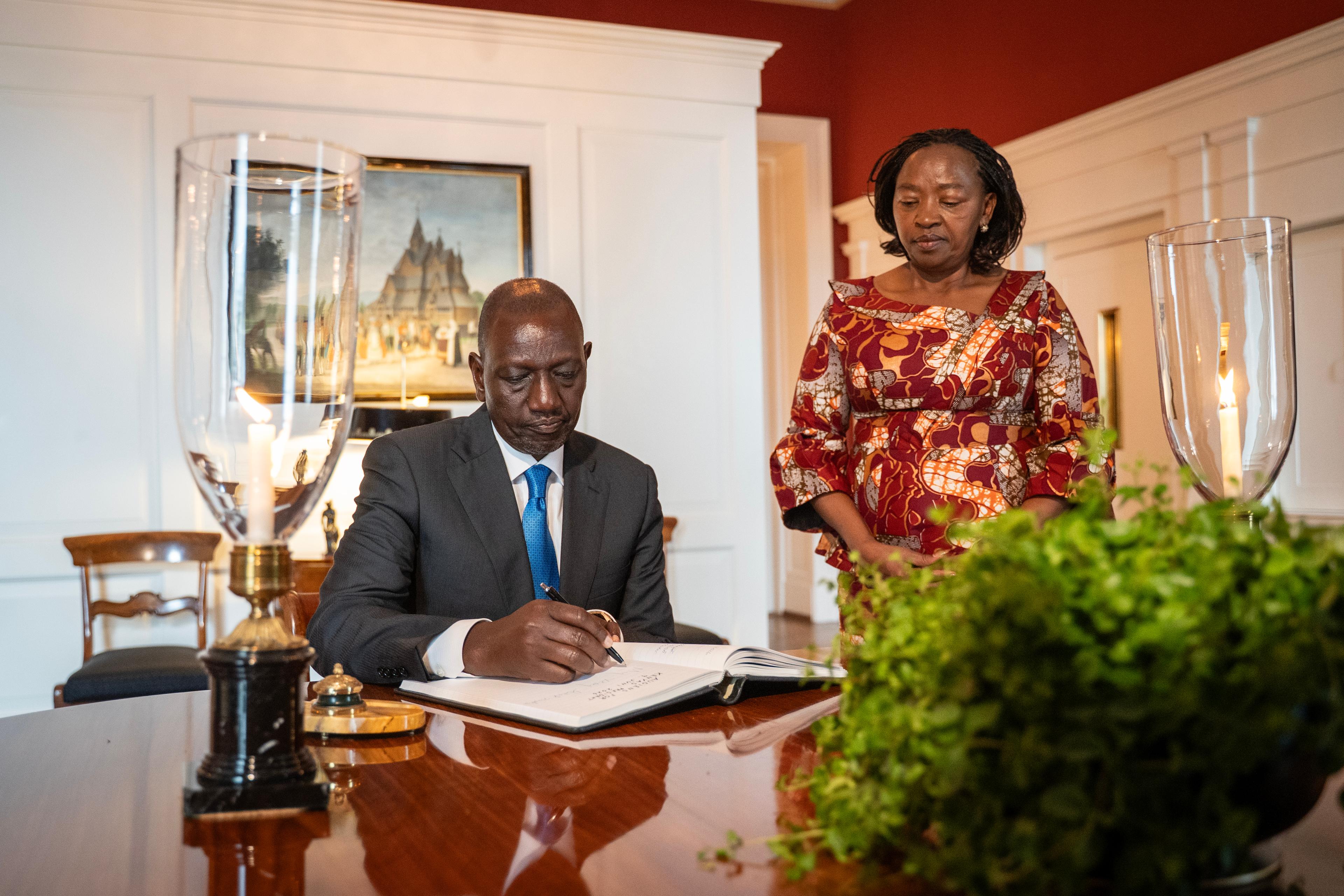 Kenyas president sitter ved et bord og signerer med en penn. Kenyas førstedame står ved siden av han og ser ned i protokollen.