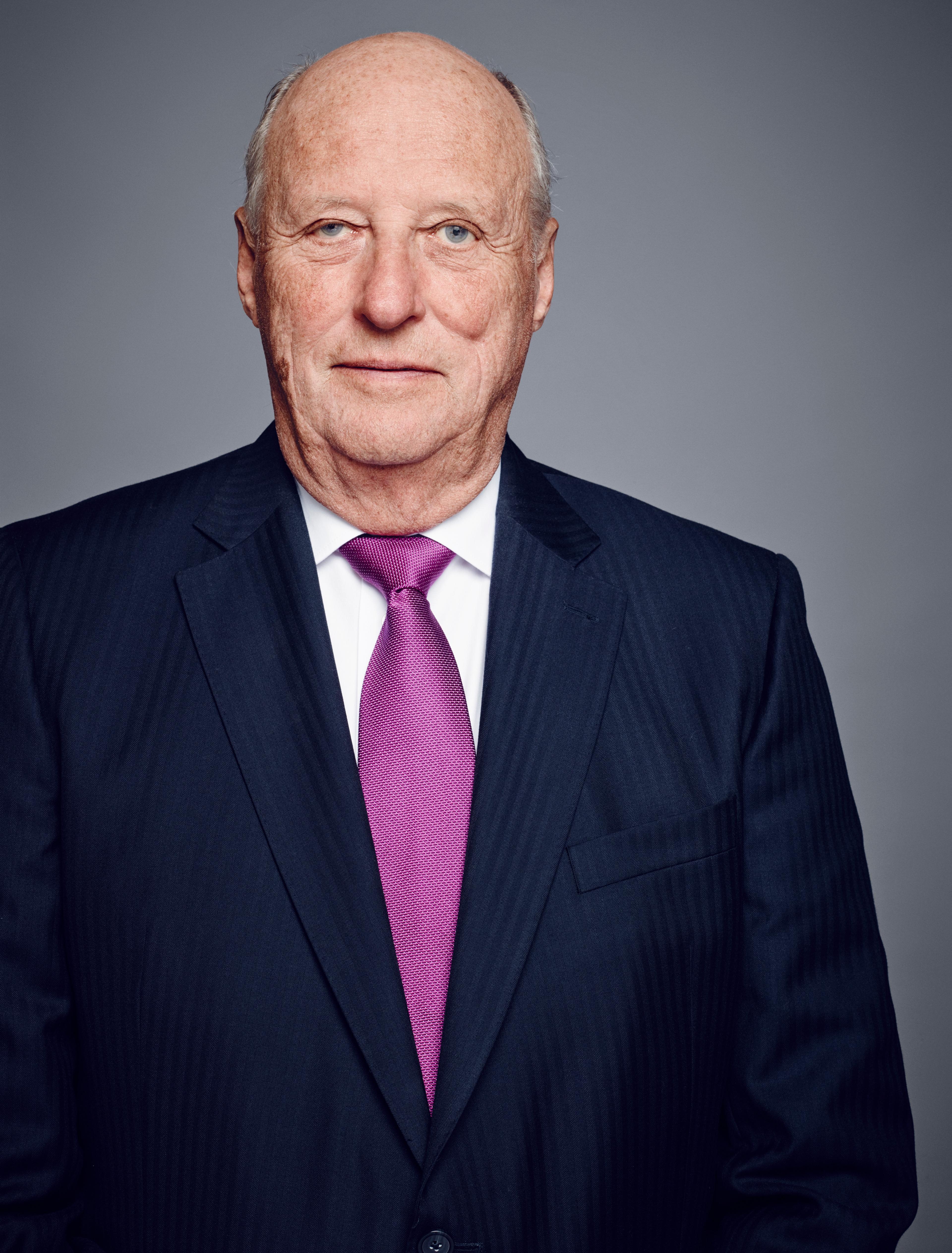 Portrett av Kong Harald i halvfigur. Kongen har mørk dress, hvit skjorte og lilla slips.