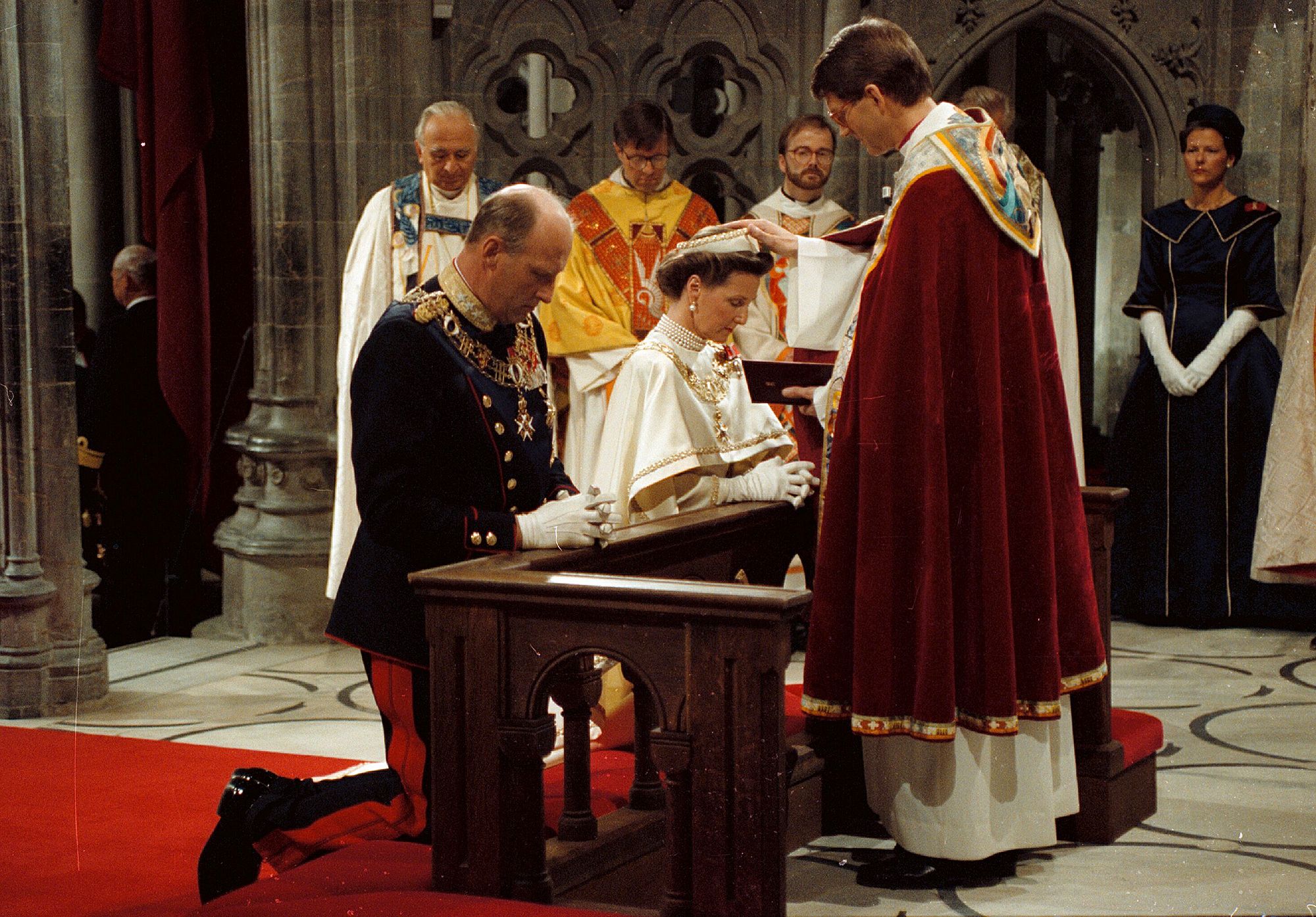 Kongeparet kneler foran biskopen, som holder hånden på Dronningens hode. Biskopen har rød kappe, Kongen er i gallauniform, Dronning Sonja en hvit kroningskjole.