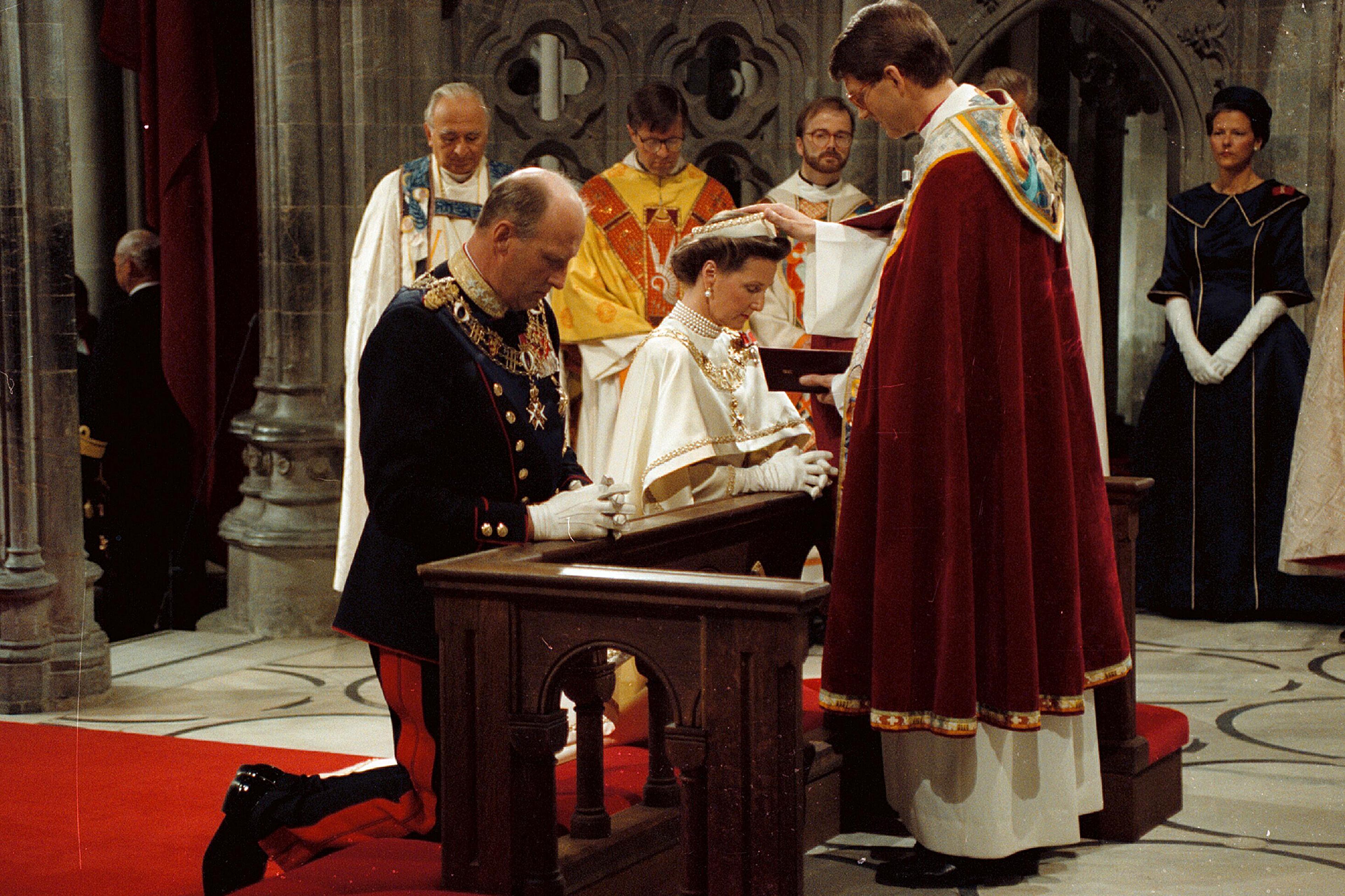 Kongeparet kneler foran biskopen, som holder hånden på Dronningens hode. Biskopen har rød kappe, Kongen er i gallauniform, Dronning Sonja en hvit kroningskjole.