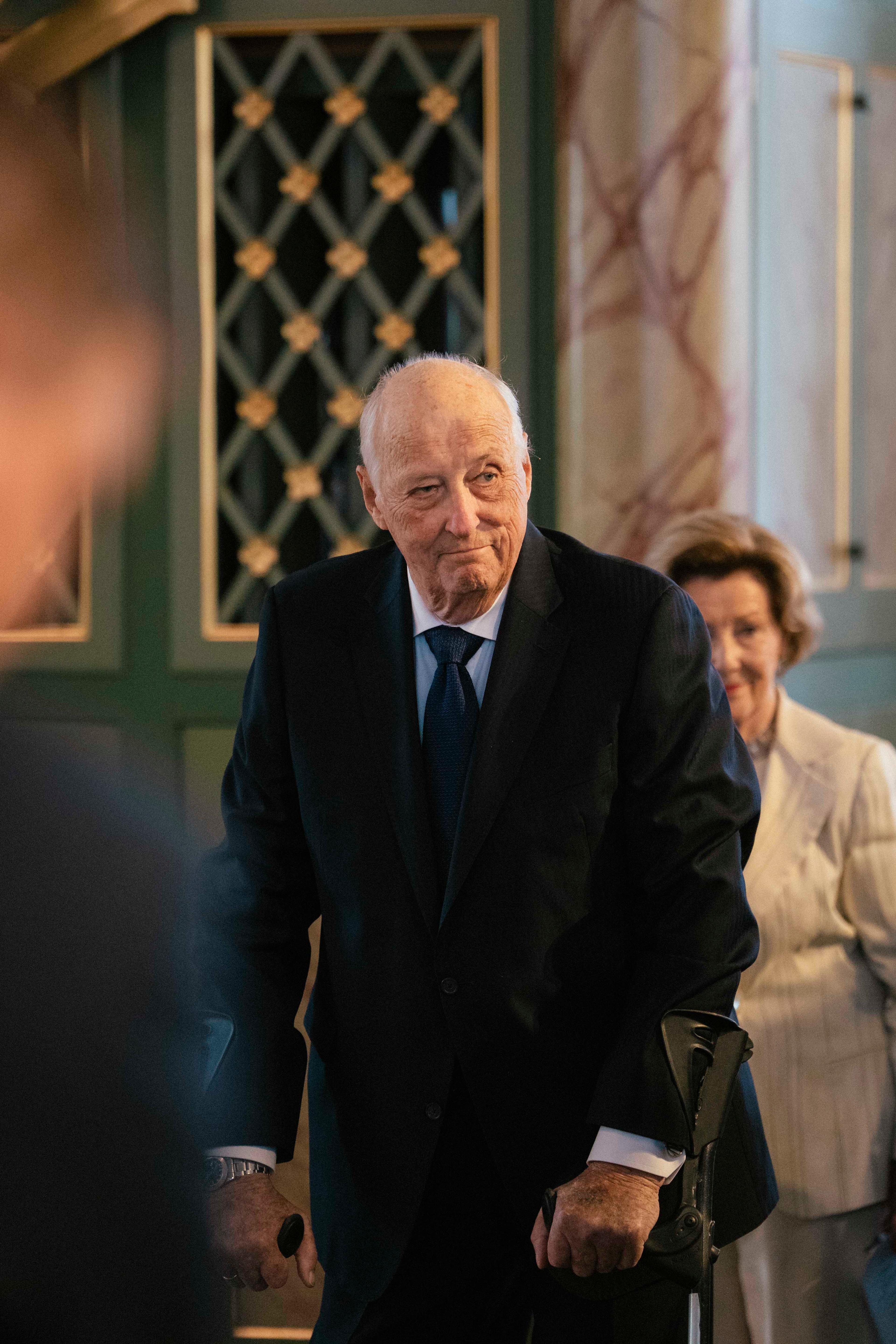 Kong Harald står oppreist ved kirkebenken. I bakgrunnen ser vi Dronning Sonja