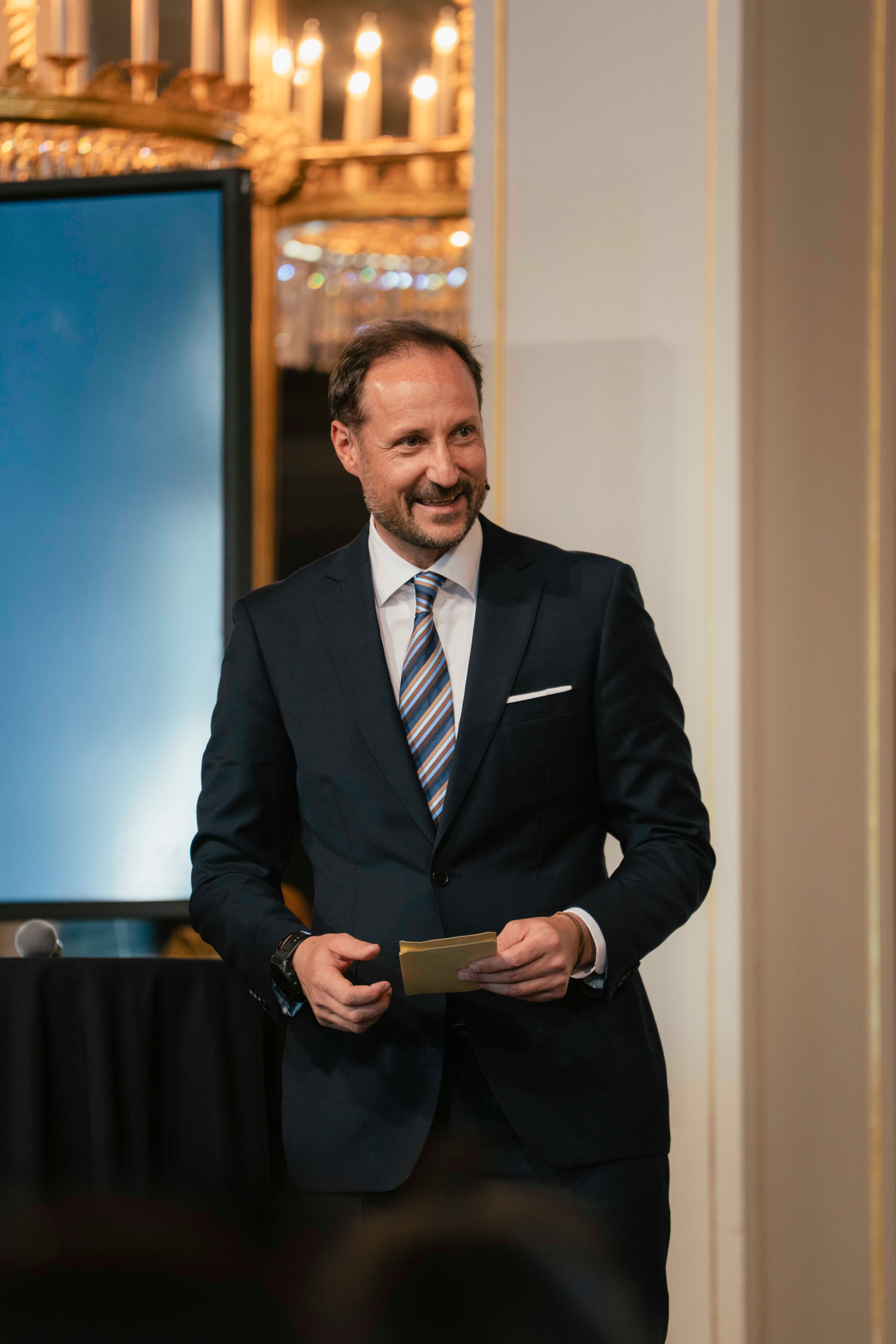 Kronprins Haakon i mørk dress og et stort smil holder talekort i hendene.