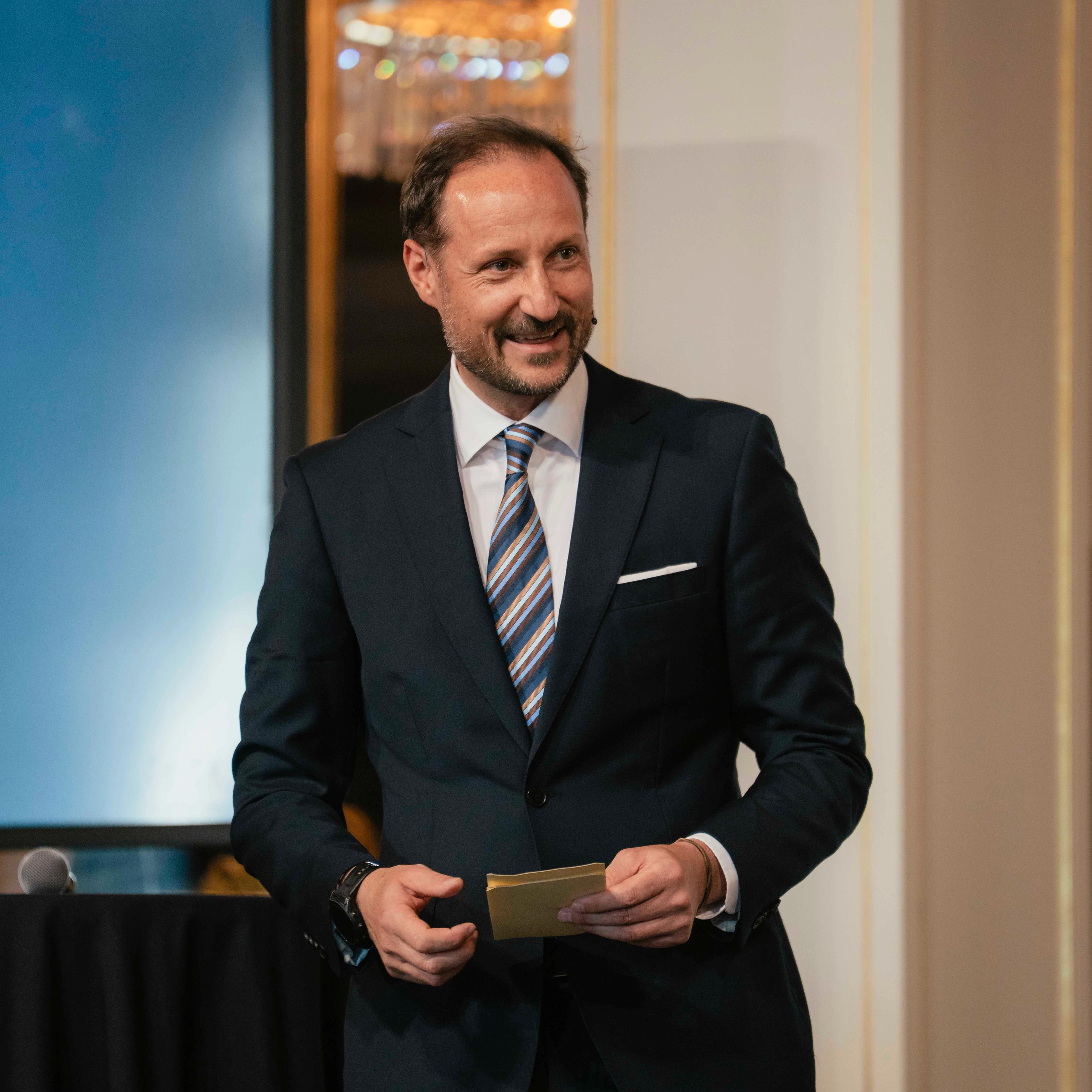 Kronprins Haakon i mørk dress og et stort smil holder talekort i hendene.