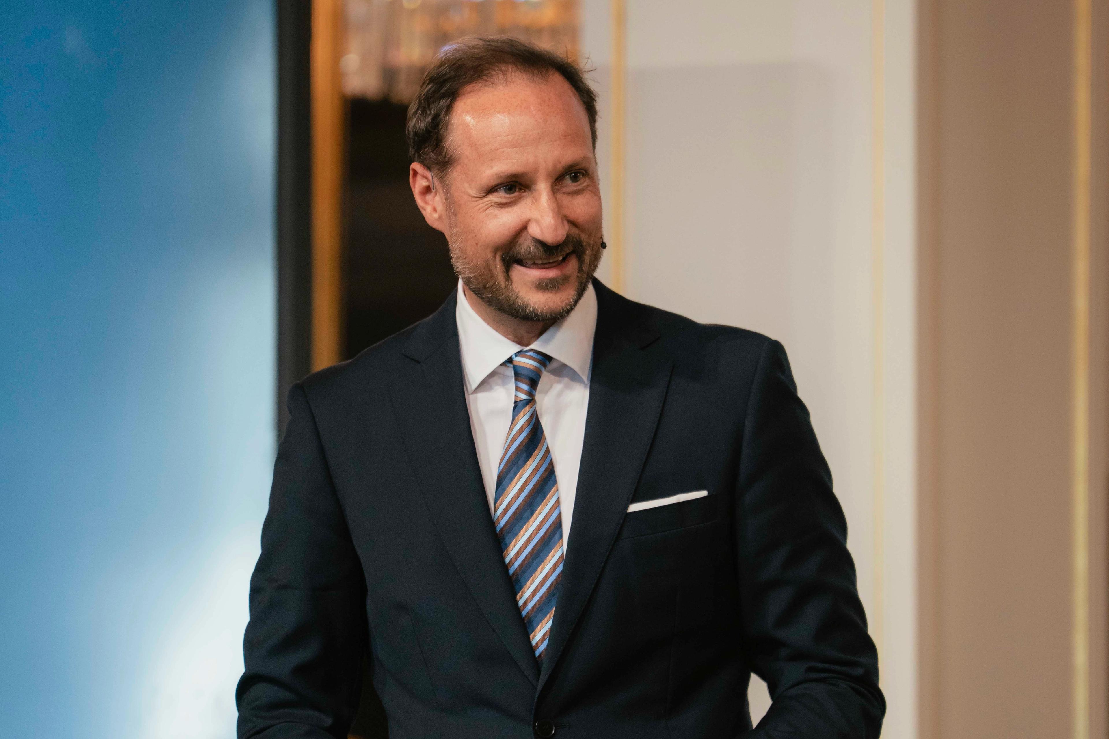 Kronprins Haakon i mørk dress og et stort smil holder talekort i hendene.