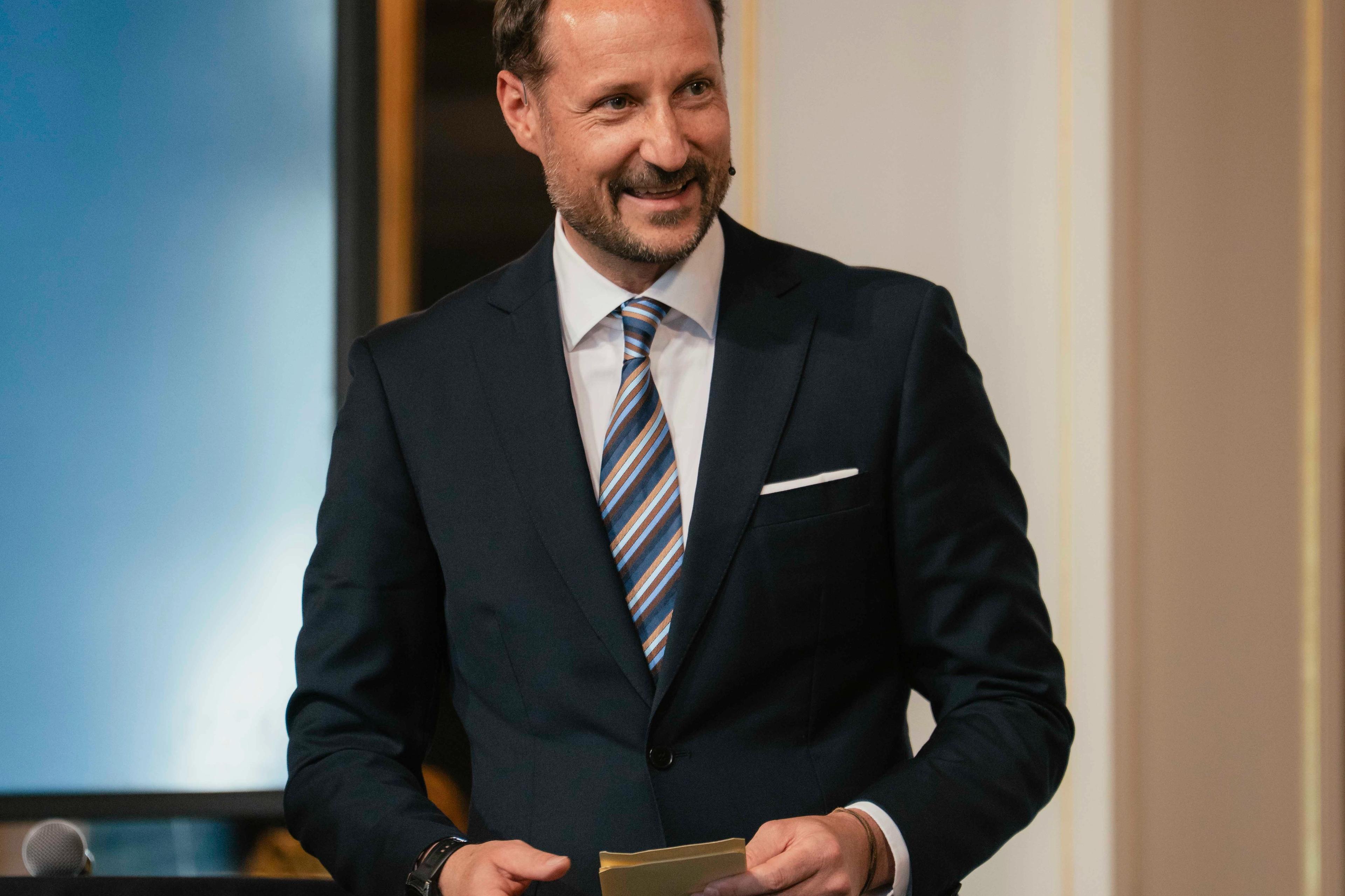 Kronprins Haakon i mørk dress og et stort smil holder talekort i hendene.
