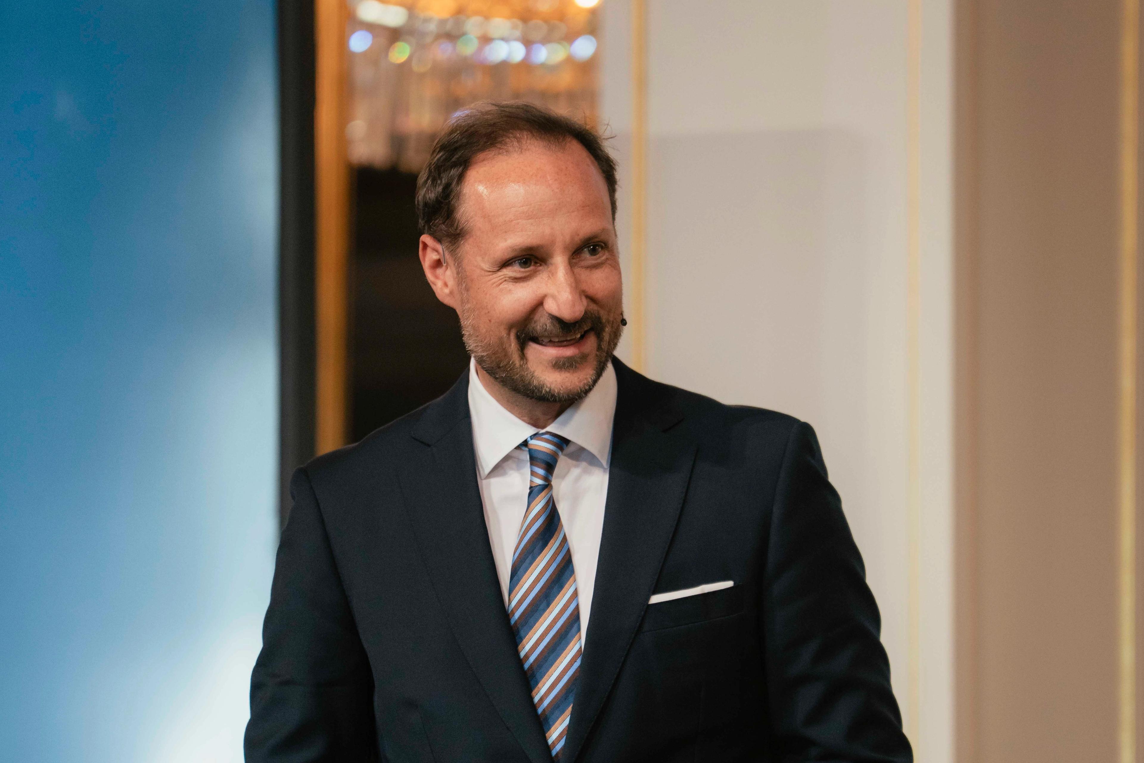 Kronprins Haakon i mørk dress og et stort smil holder talekort i hendene.