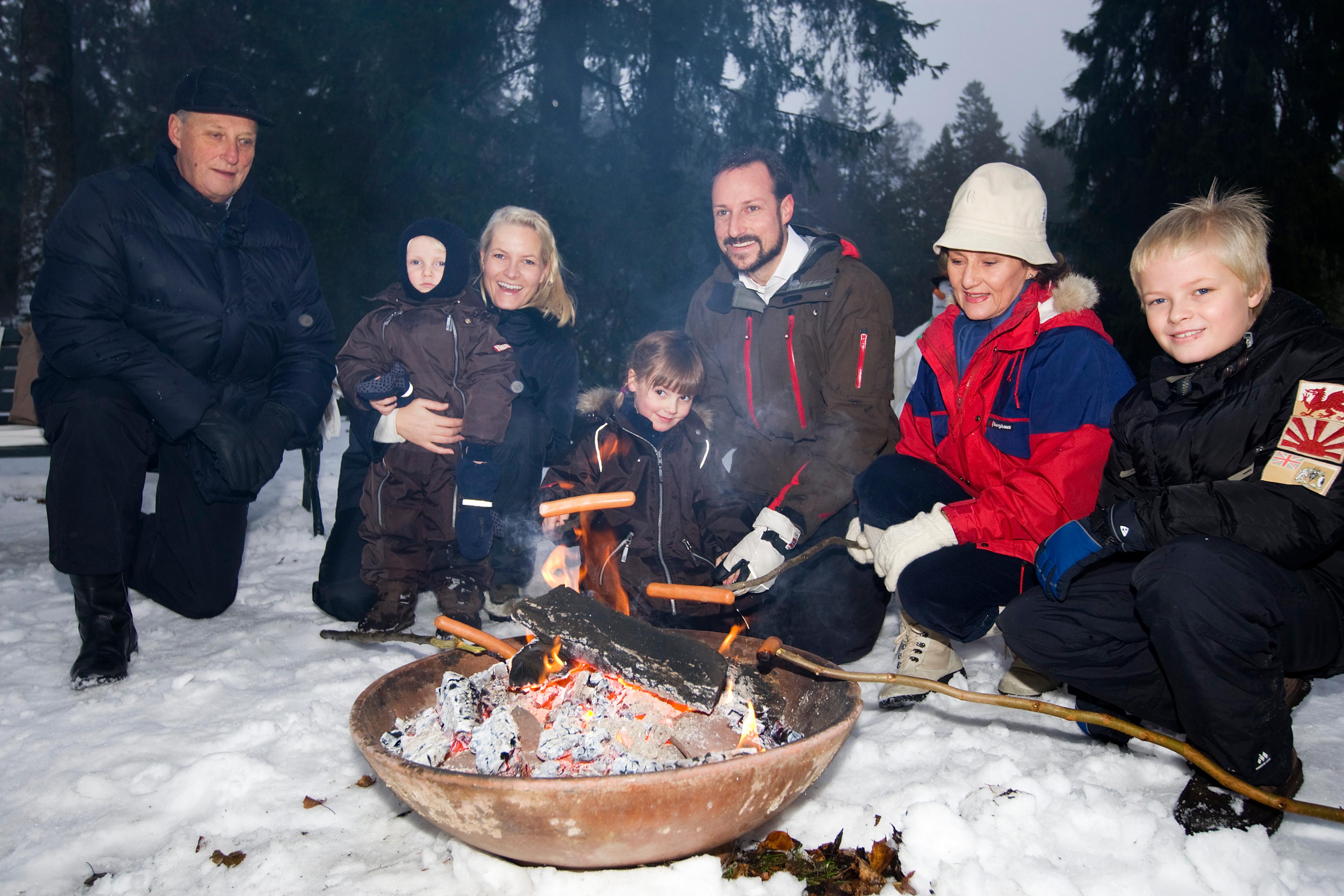 Kong Harald, Dronning Sonja og Kronprinsfamilien steker pølse på bålbanne i skogen.
