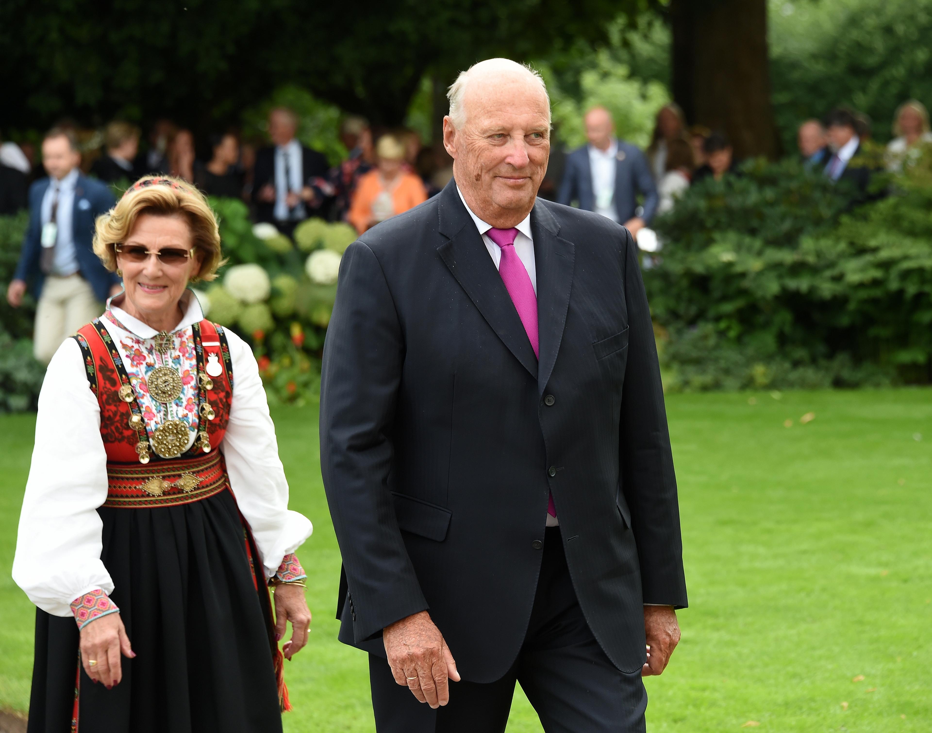 Kongen og Dronningen går sammen i Slottsparken. Dronningen er i bunad, Kongen i mørk dress og lilla slips. Det er mange mennesker i bakgrunnen.