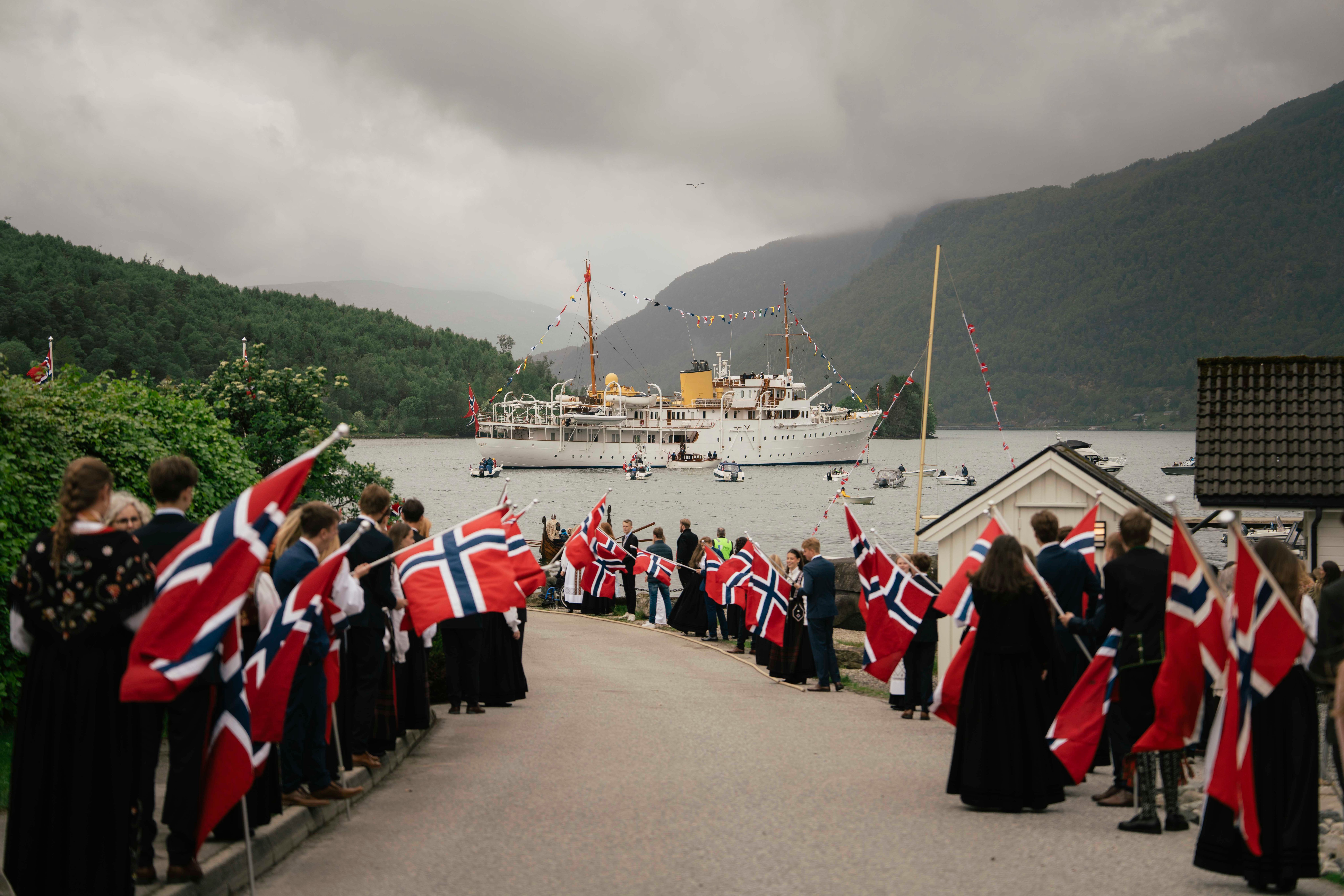 Elevar står langs vegen med store flagg. I fjorden ligg Kongeskipet.