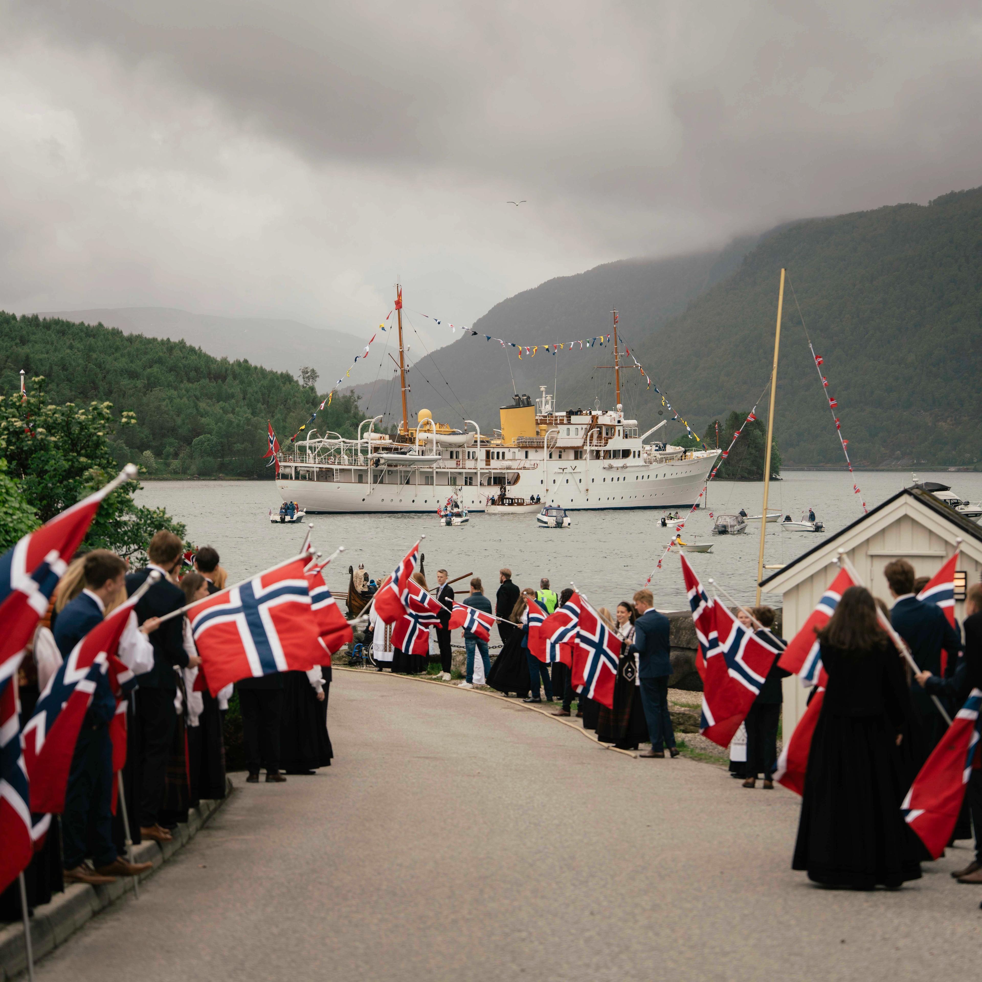 Elevar står langs vegen med store flagg. I fjorden ligg Kongeskipet.