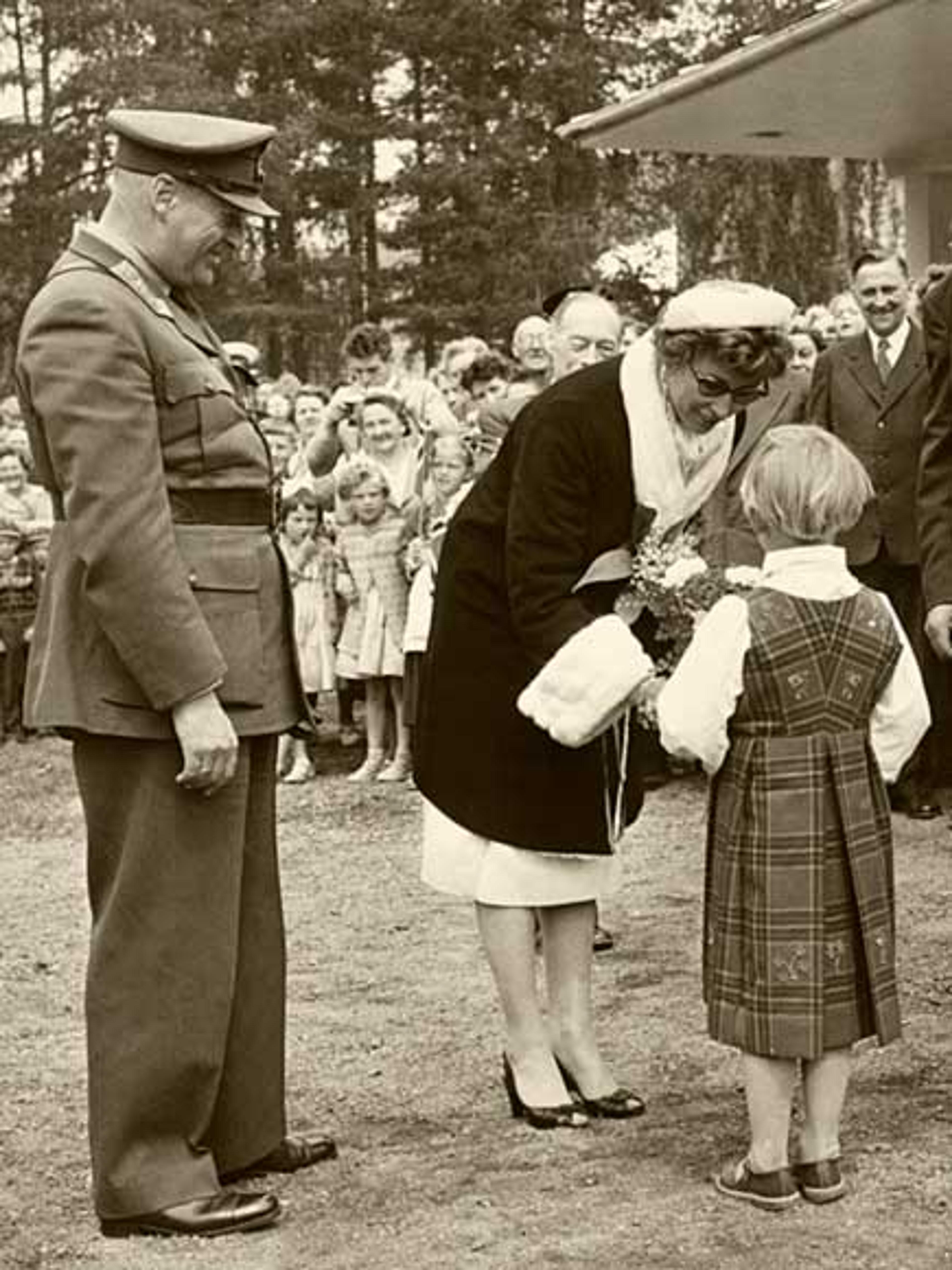 Sort-hvittbilde av Prinsesse Astrid som bøyer seg ned og får en blomsterbukett av en liten pike. Kong Olav står smilende ved siden av og ser på.