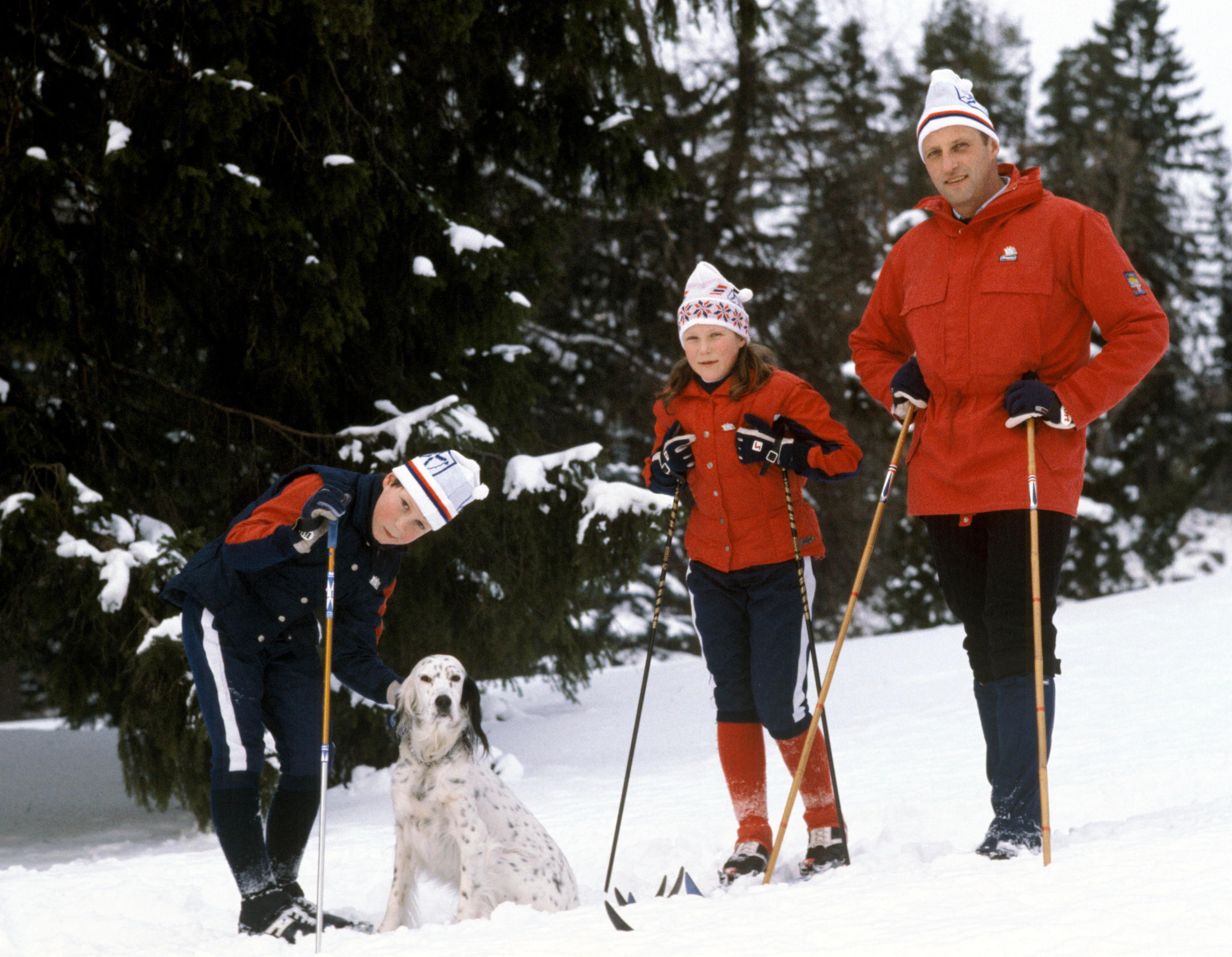 Kronprins Harald står i skiutstyr sammen med barna, Prinsesse Märtha Louise og Prins Haakon, som klapper familiens hund.