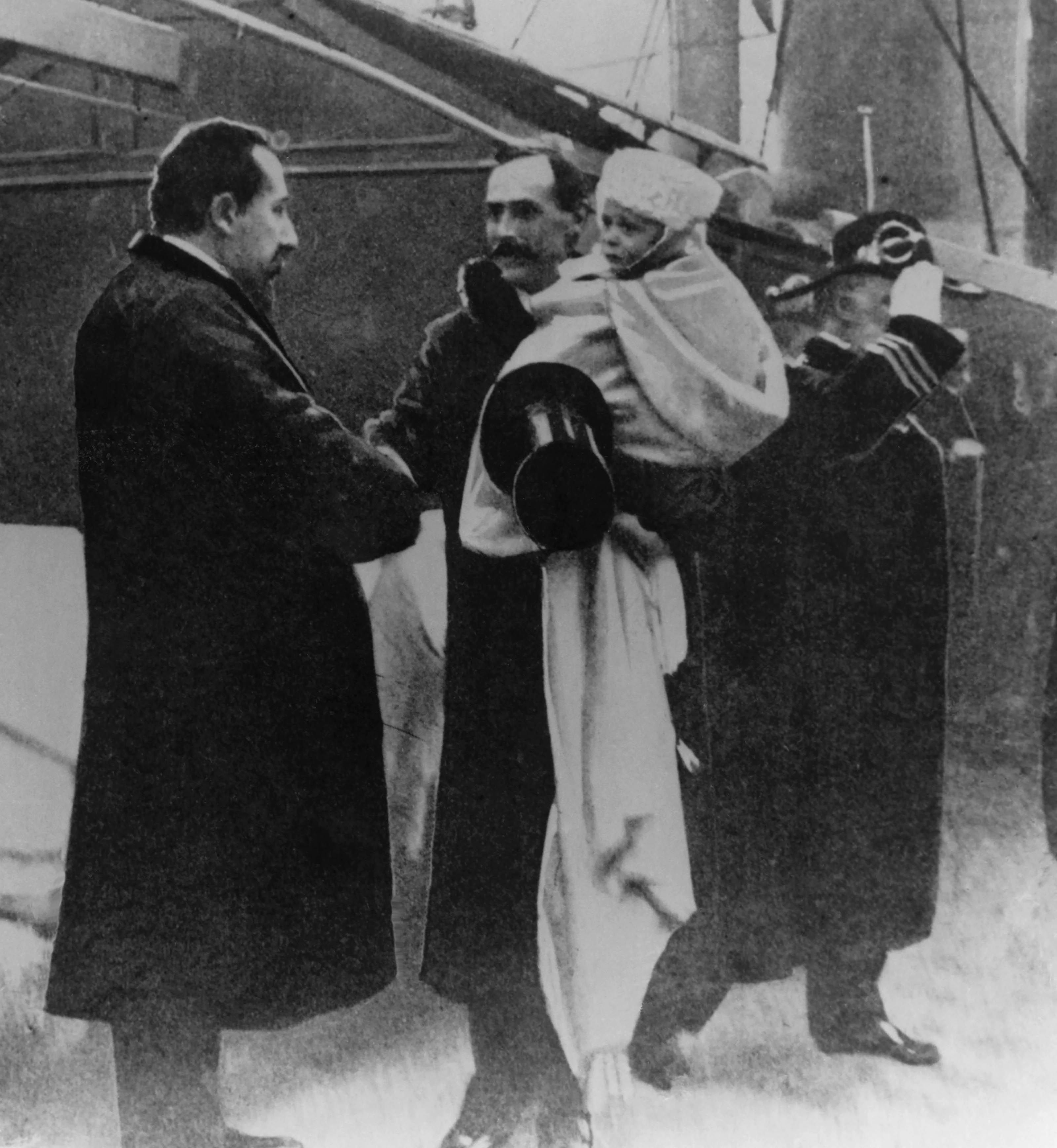Sort-hvitt bilde av møtet mellom Kong Haakon og statsministeren på kaia i Oslo, november 1905. To år gamle Kronprins Olav sitter innhyllet i pledd på farens arm. 