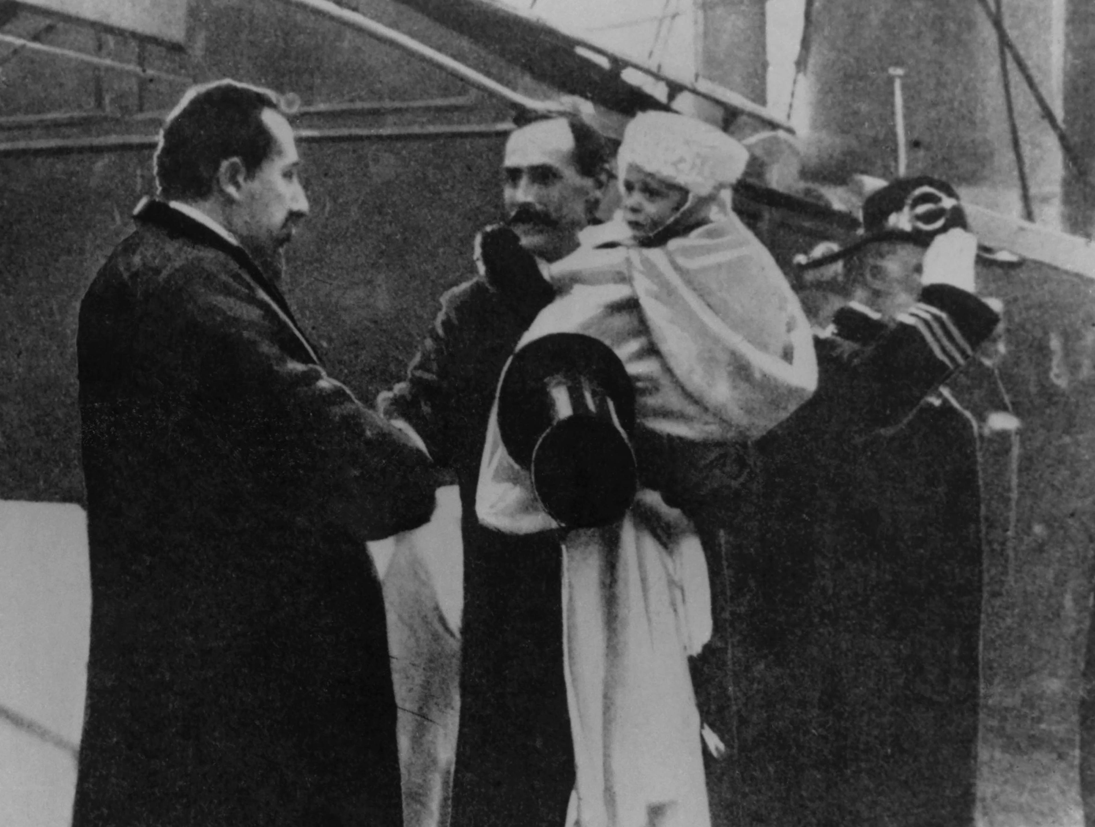 Sort-hvitt bilde av møtet mellom Kong Haakon og statsministeren på kaia i Oslo, november 1905. To år gamle Kronprins Olav sitter innhyllet i pledd på farens arm.