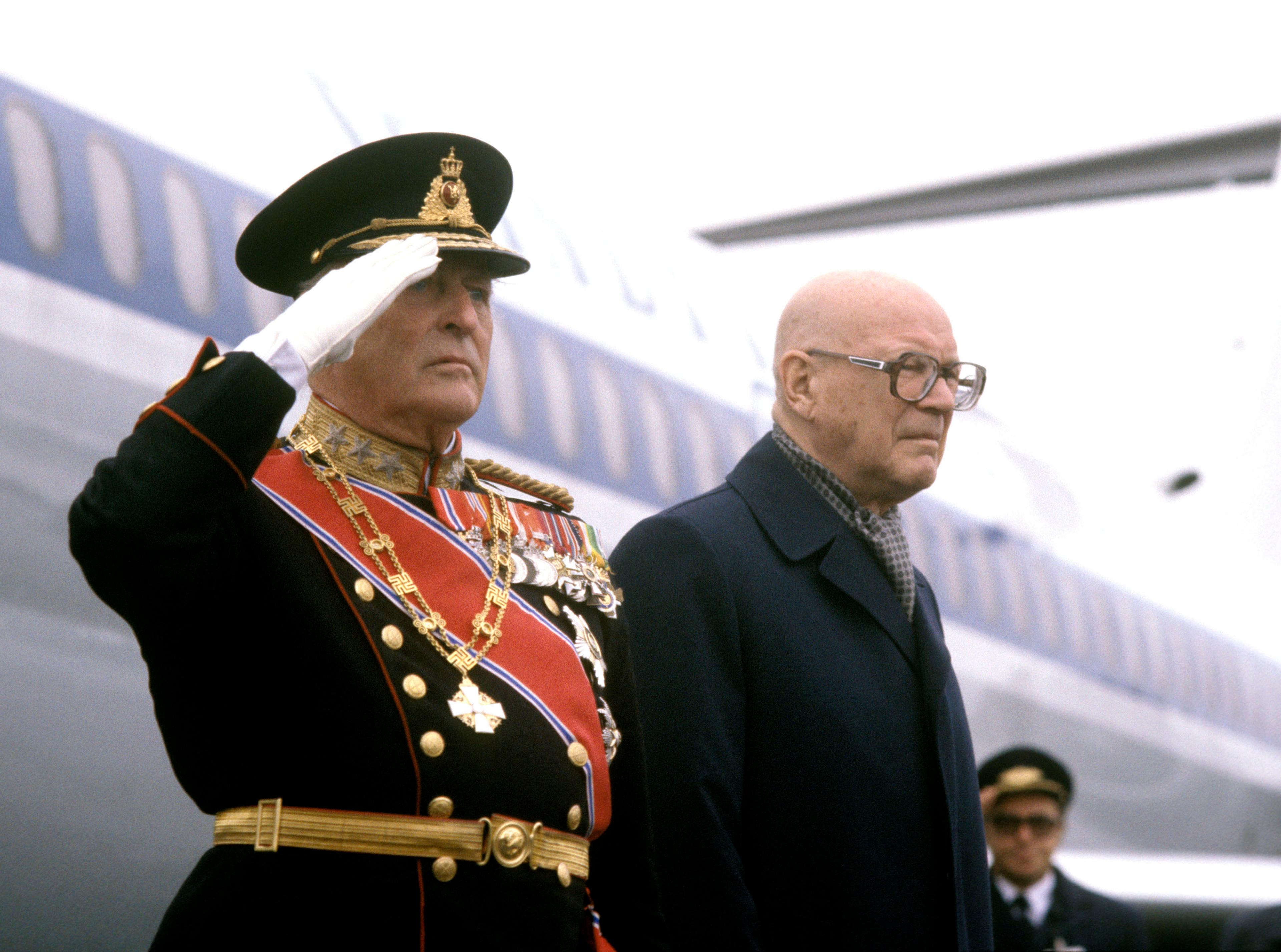 Kong Olav i gallauniform gjør honnør utenfor flyet.