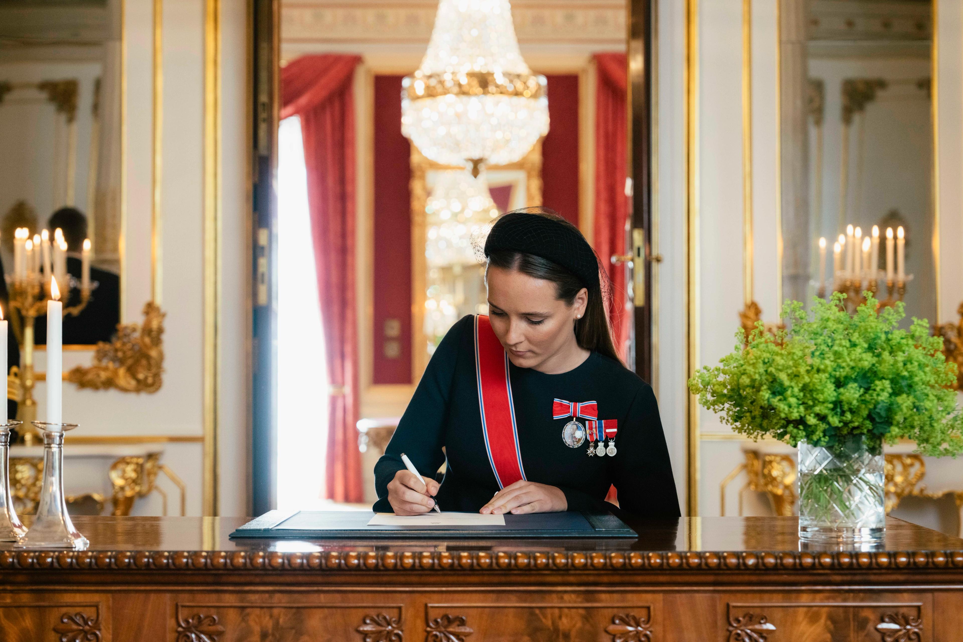 Kronprinsessen sitter bak et gedigent skrivebord og signerer. Hun er kledd i sort kjole og ordener.