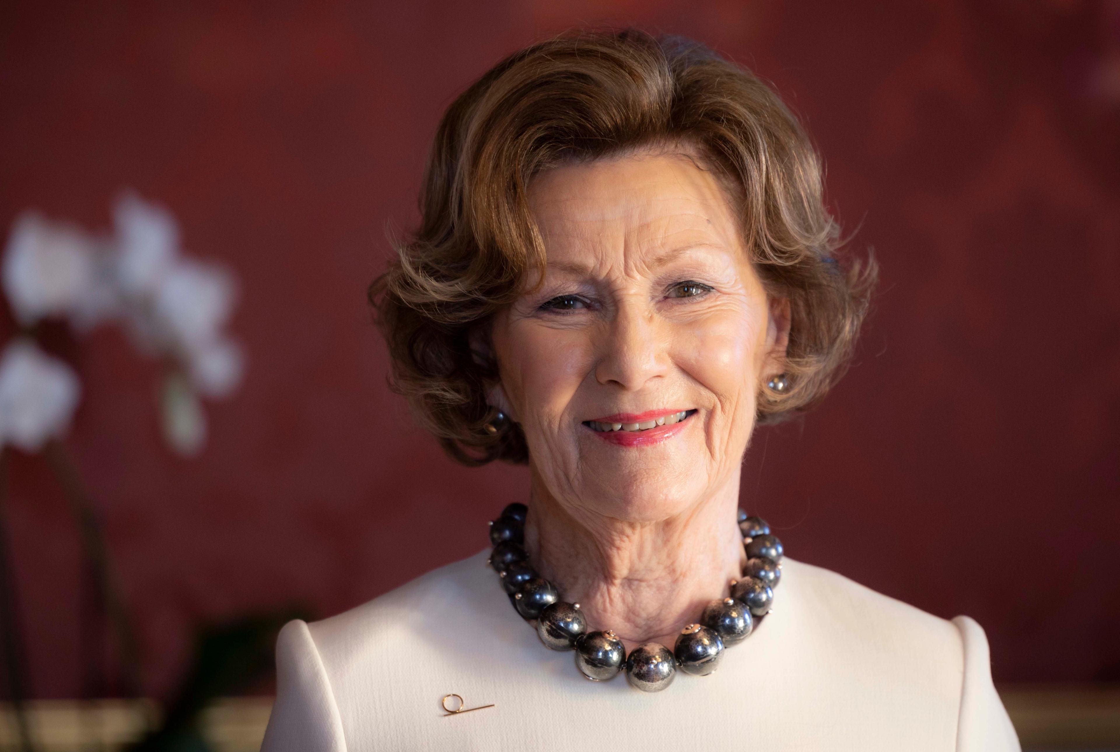 Næbilde av en smilende Dronning Sonja i en hvit kjole og kuleformetkjede rundt halsen. Bakgrunnen er dyp rød, med et hint av noen orkideer på venstre side.