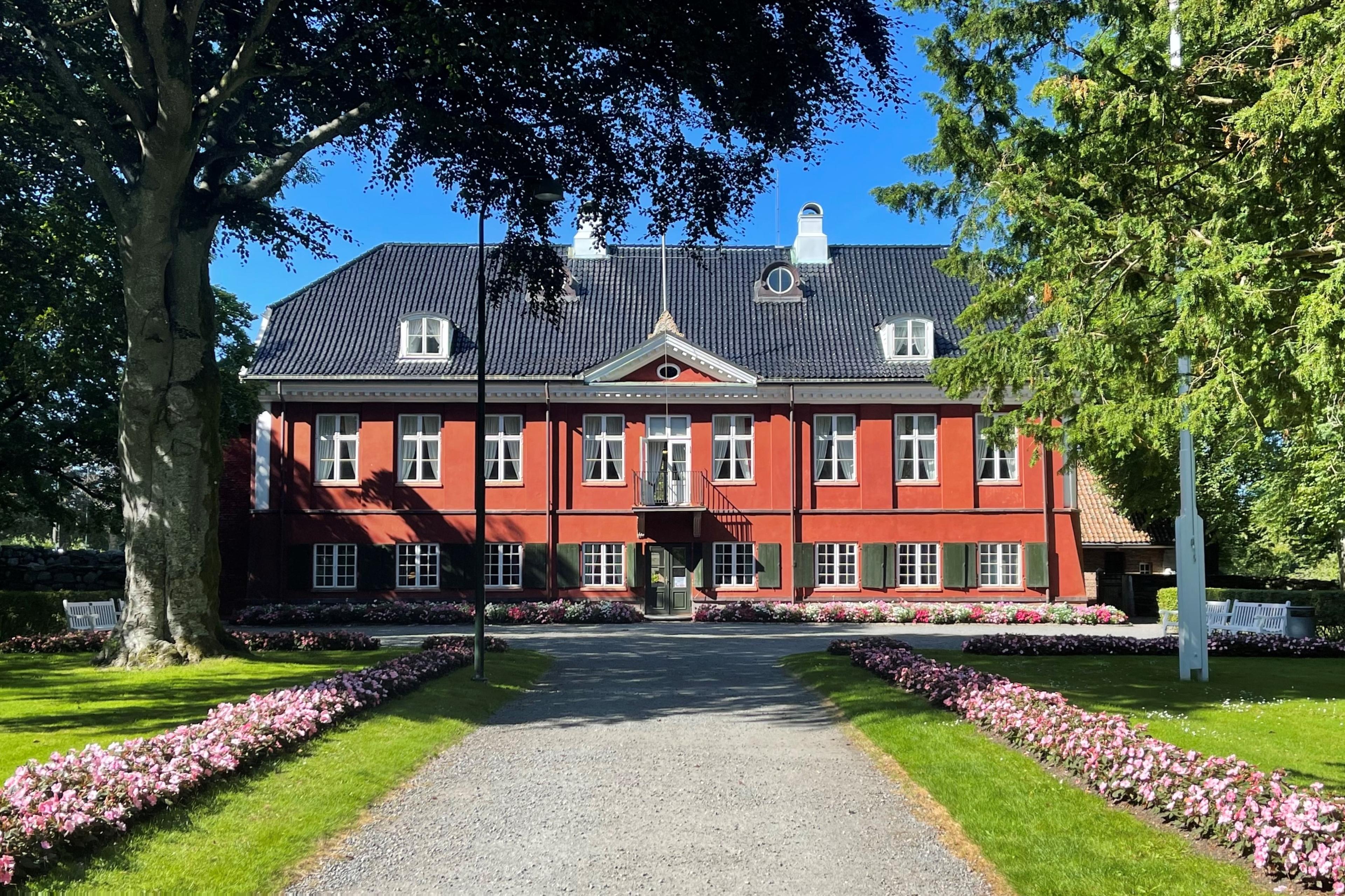 En flott treetasjes bygning i rød teglstein ligger omkranset av en park for enden av en grusvei.