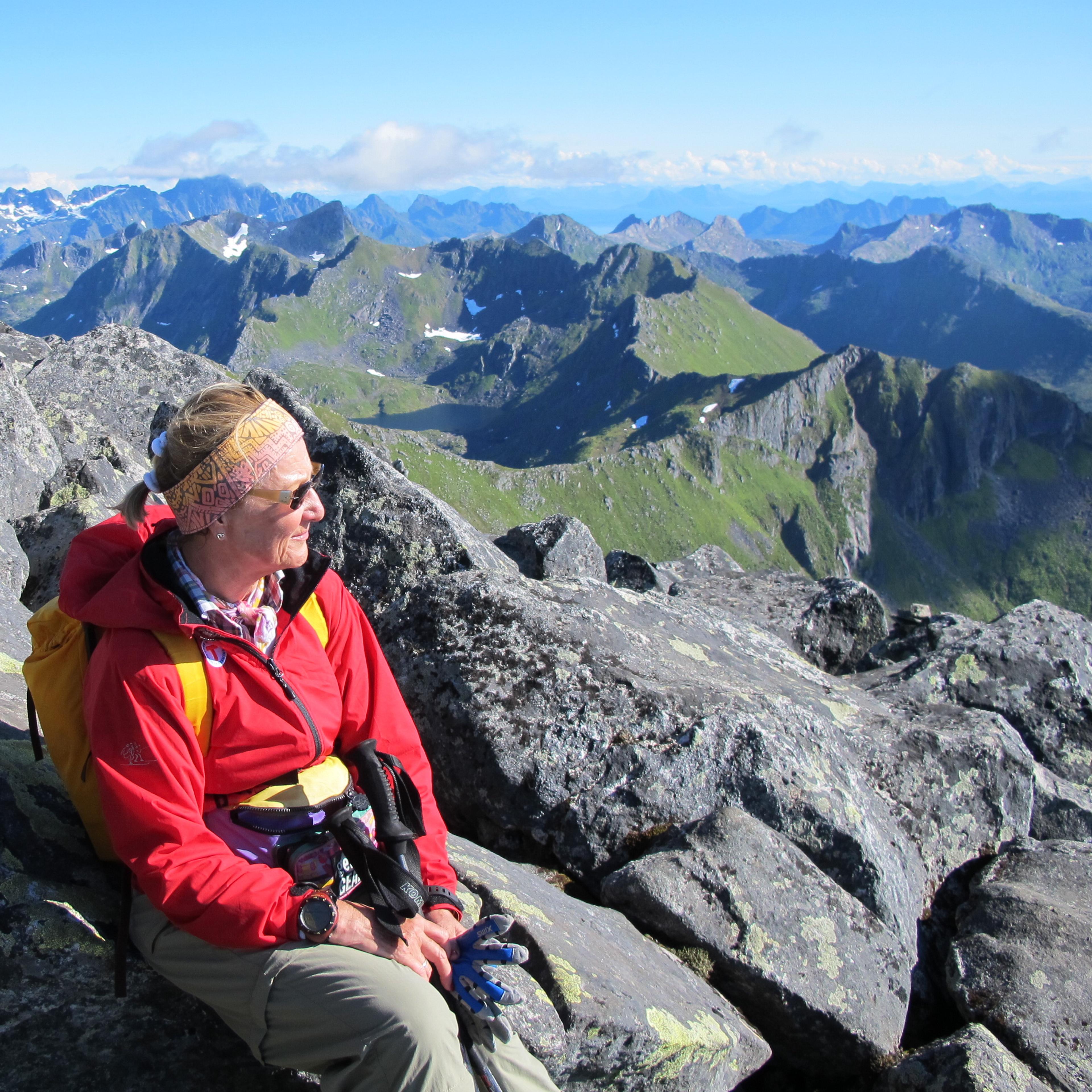 Dronningen sitter på en sten og ser ut over fjellandskapet. Hun er kledd i turklær, med rød anorakk og solbriller.