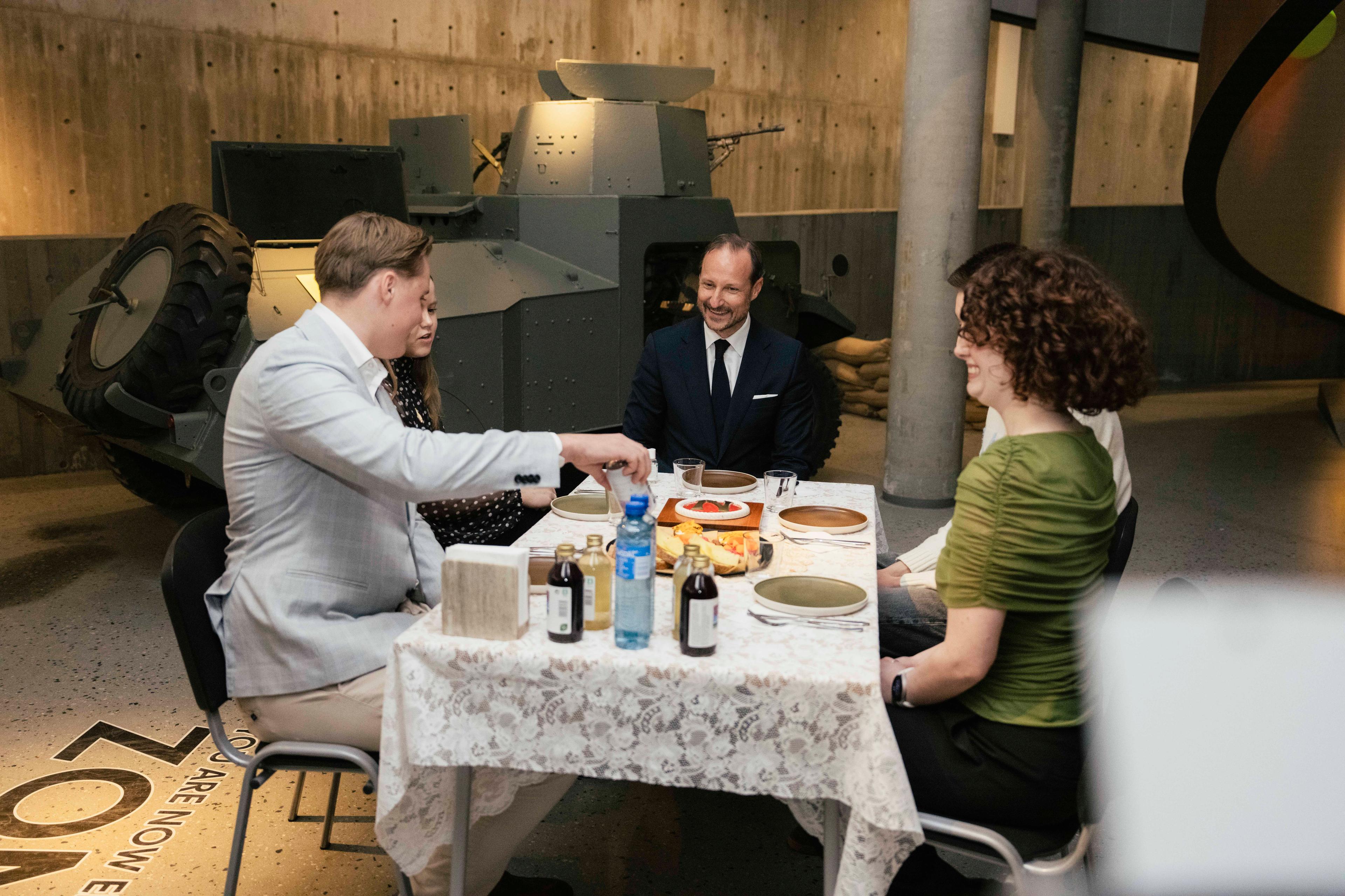 Kongen og fire ungdommer sitter ved et dekket bord i et rom fylt med krigsmateriell.