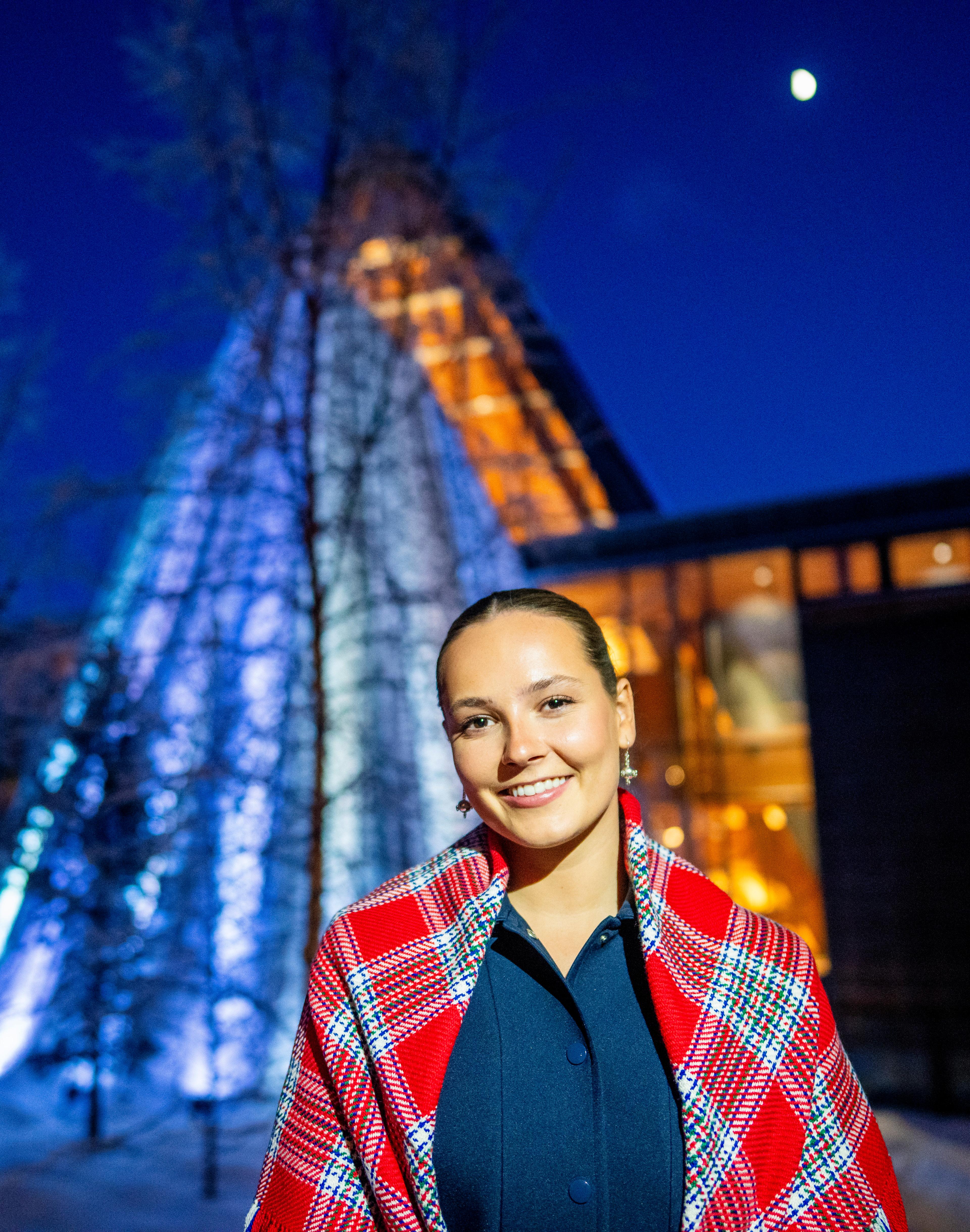Tett på Prinsesse Ingrid Alexandra i mørke ute, ikledd samisk sjal og med en lyssatt sametingsbygning bak.