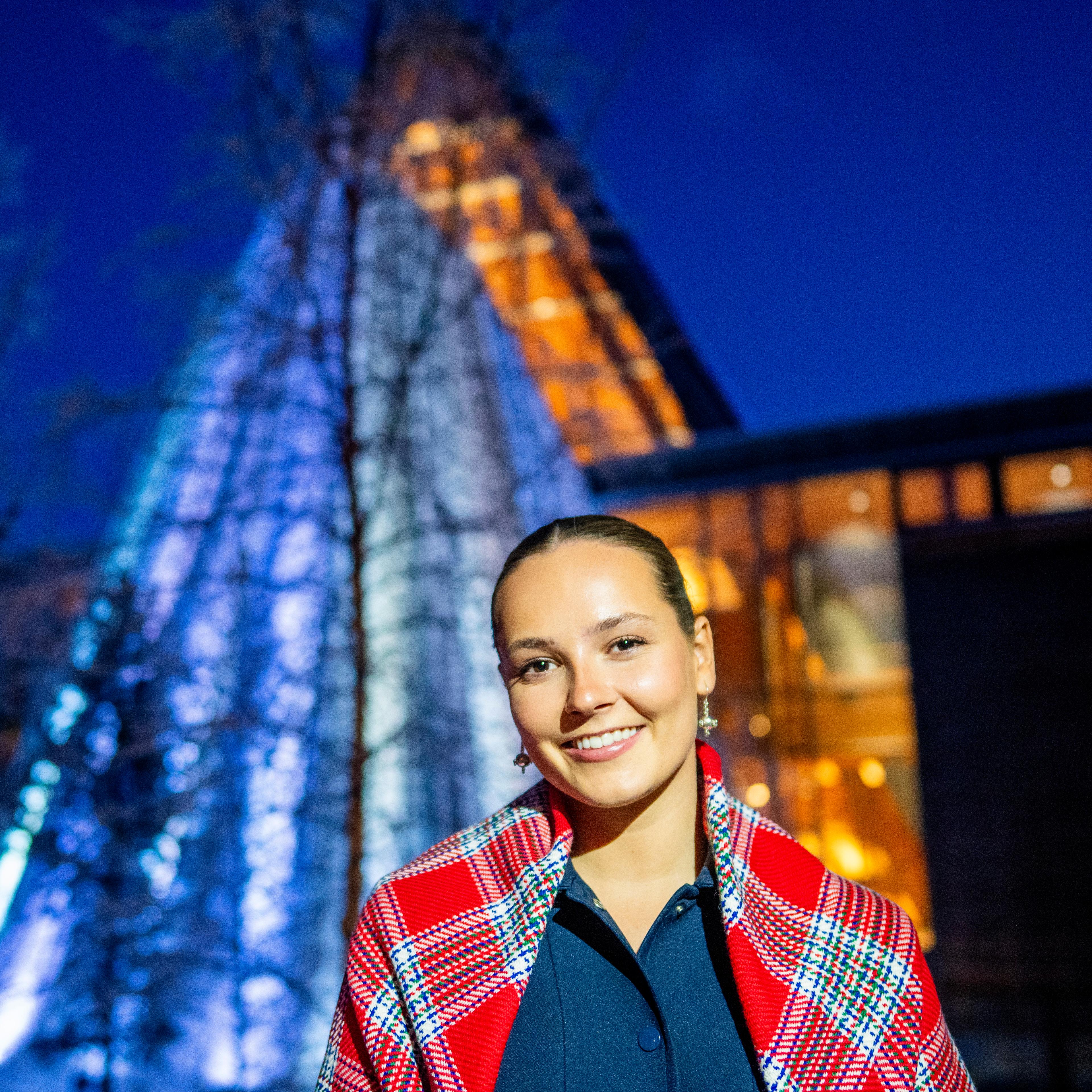 Tett på Prinsesse Ingrid Alexandra i mørke ute, ikledd samisk sjal og med en lyssatt sametingsbygning bak.